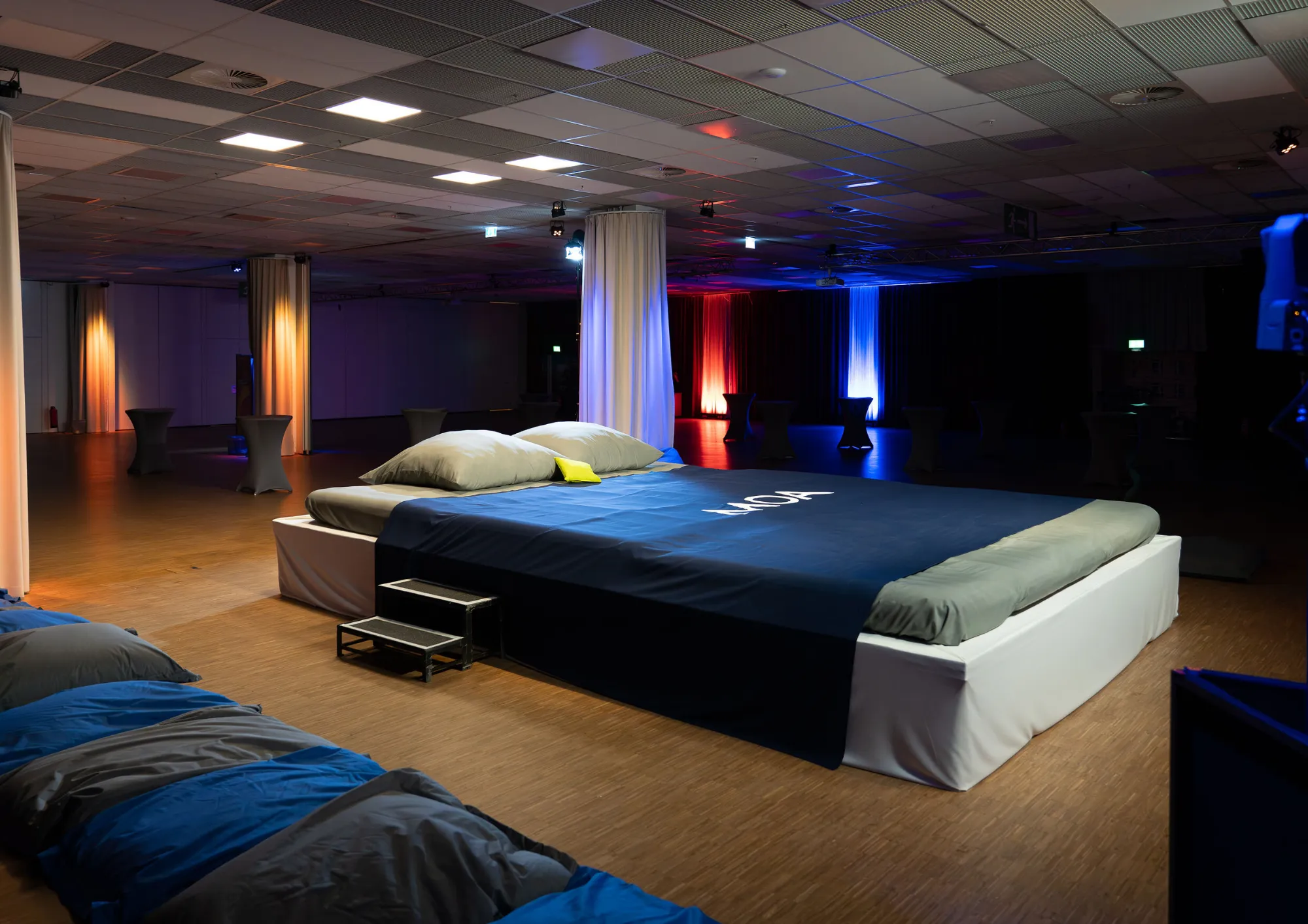 Ein großes Bett mit weißen Kissen steht in einer abgedunkelten Veranstaltungshalle mit farbiger Beleuchtung und weiteren Liegeflächen.