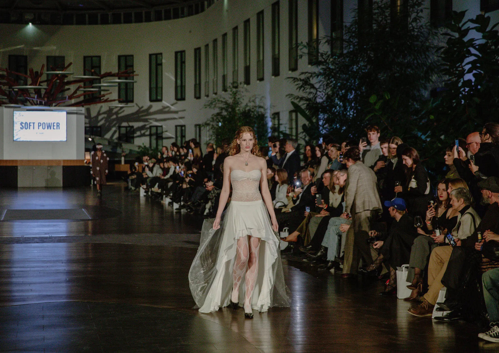 Eine Models läuft in einem hellen Kleid durch ein Atrium, während zahlreiche sitzende Gäste entlang des Laufstegs zuschauen.