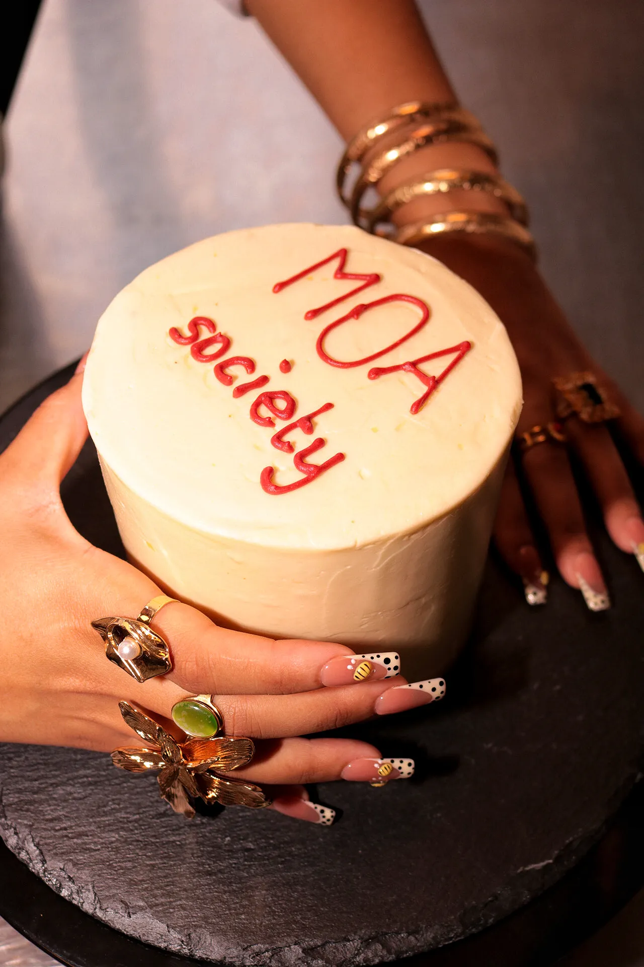 A round cake with the red lettering "MOA Society" is held by hands with striking jewellery.