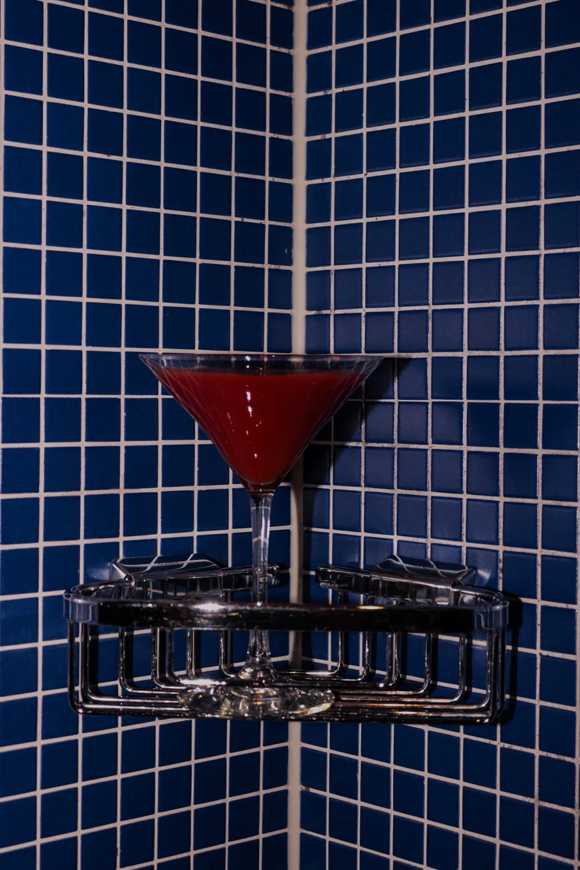 A red cocktail stands in a martini glass on a chrome wall shelf against dark blue tiles with white grout.