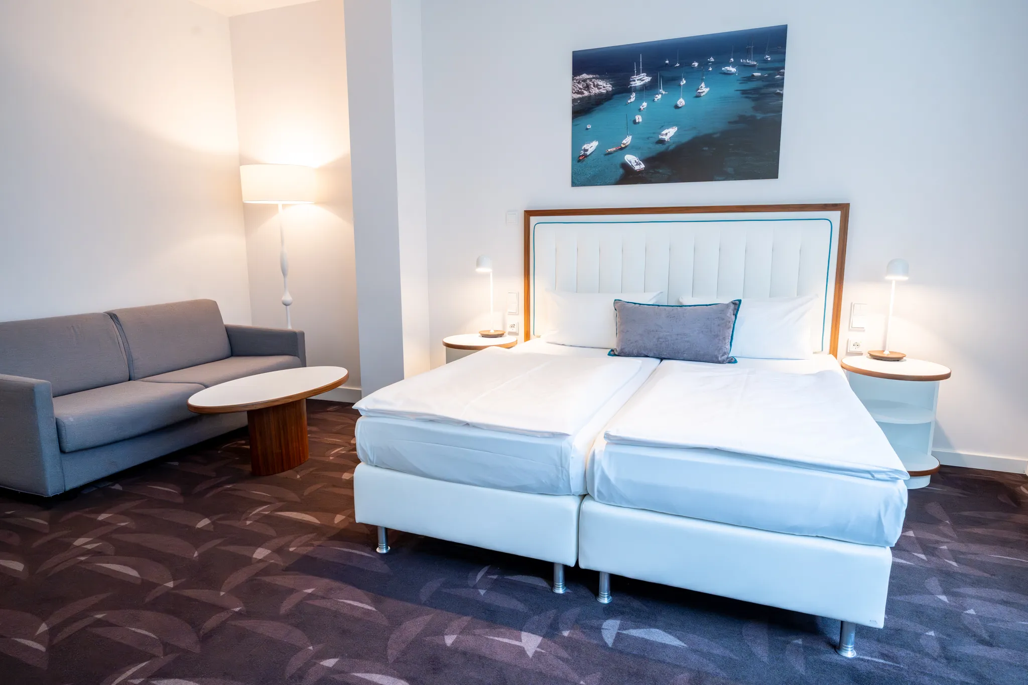 Ein Hotelzimmer mit Doppelbett, grauem Sofa, rundem Beistelltisch, Nachttischen, Stehlampen und einem gerahmten Bild an der Wand.