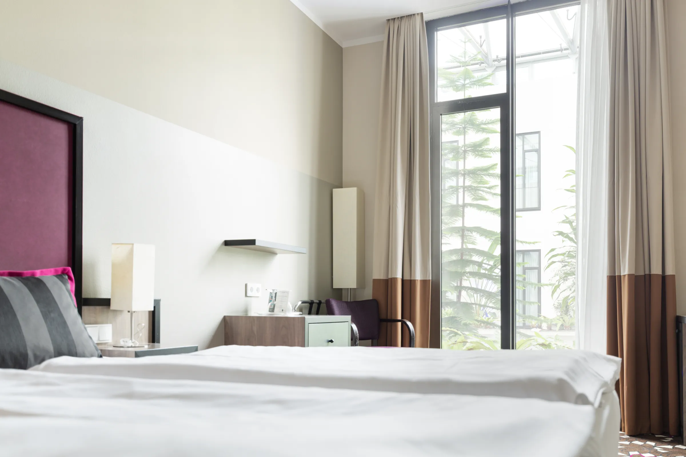Helles Hotelzimmer mit Doppelbett, Schreibtisch, Stuhl, Küchenzeile und bodentiefen Fenstern mit Vorhängen.