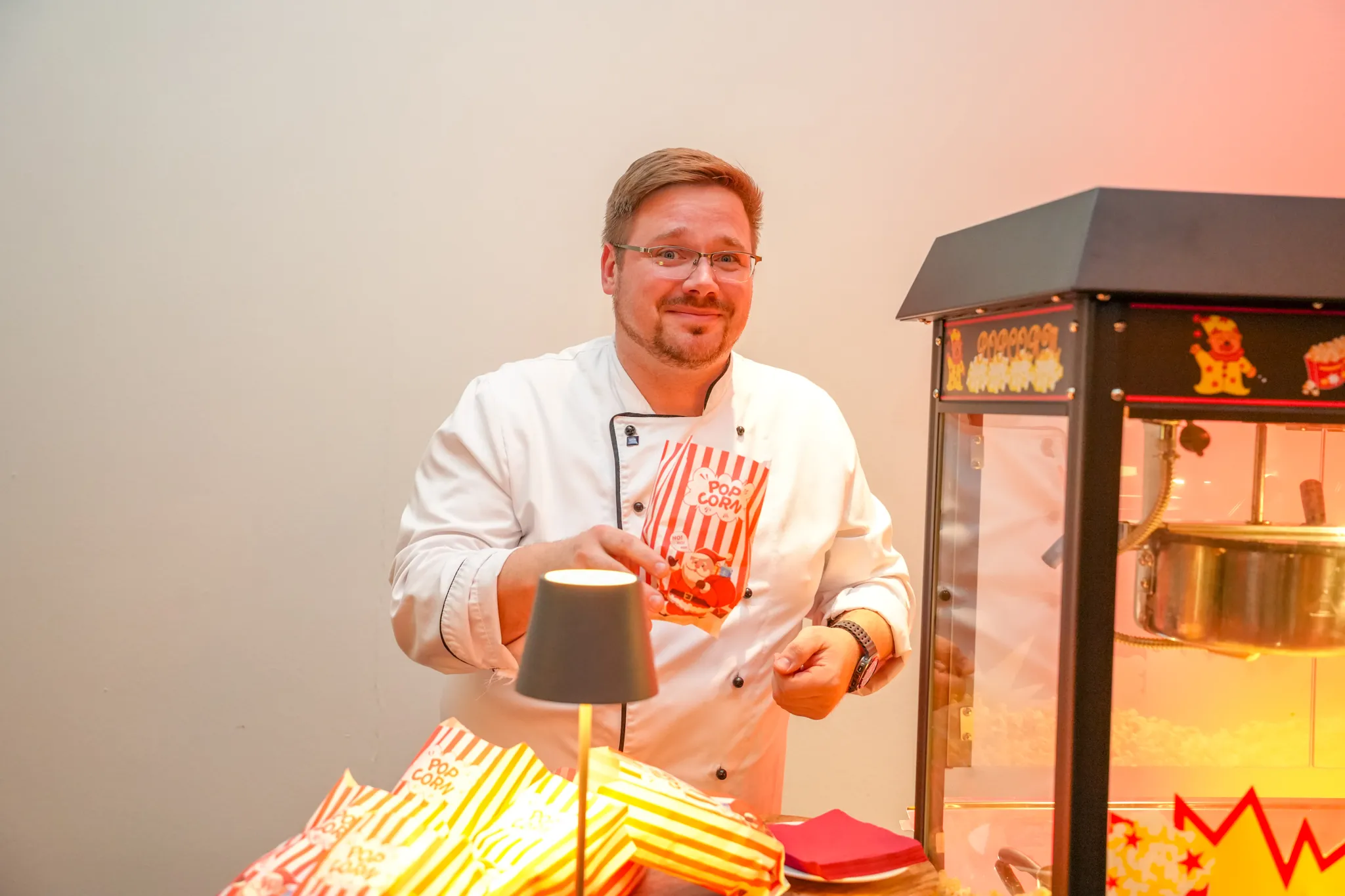 Ein Koch steht neben einer beleuchteten Popcornmaschine und hält eine Schale mit Popcorn in den Händen.