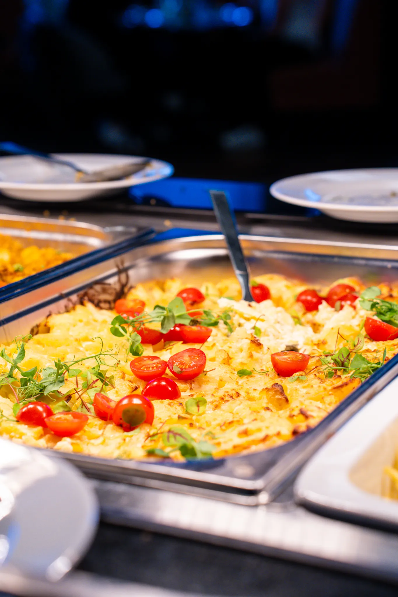 Ein Buffet mit einer goldbraun überbackenen Speise, Kirschtomaten und Kräutern in einer Edelstahlwanne.