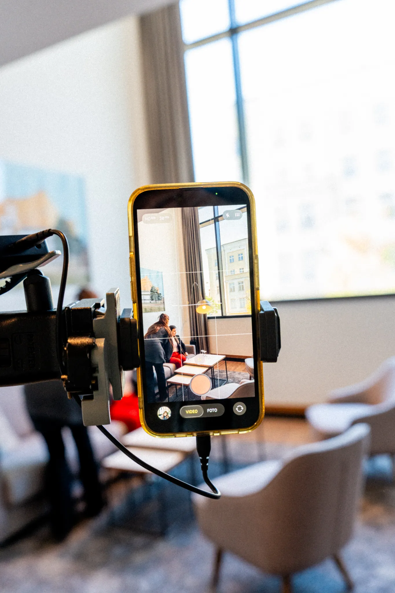 A smartphone on a camera tripod shows a recording of a seated person at a table in a bright room.