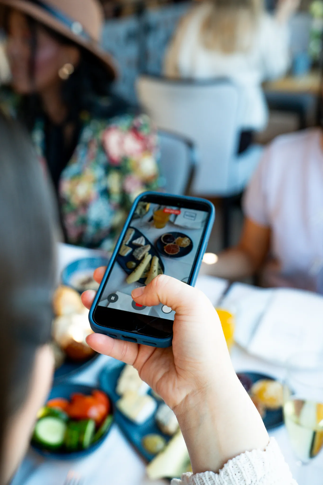A hand holds a smartphone showing a recording of a plated dish, while people sit at a set table in the background.