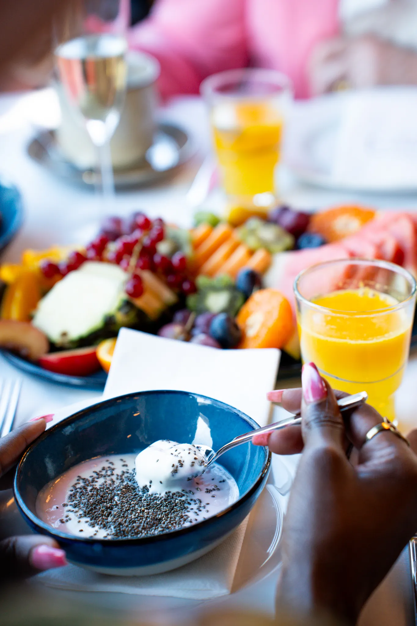 Ein Frühstückstisch mit Joghurt, Obst, Müsli, Orangensaft, Kaffee und Sekt, während eine Person mit einem Löffel umrührt.