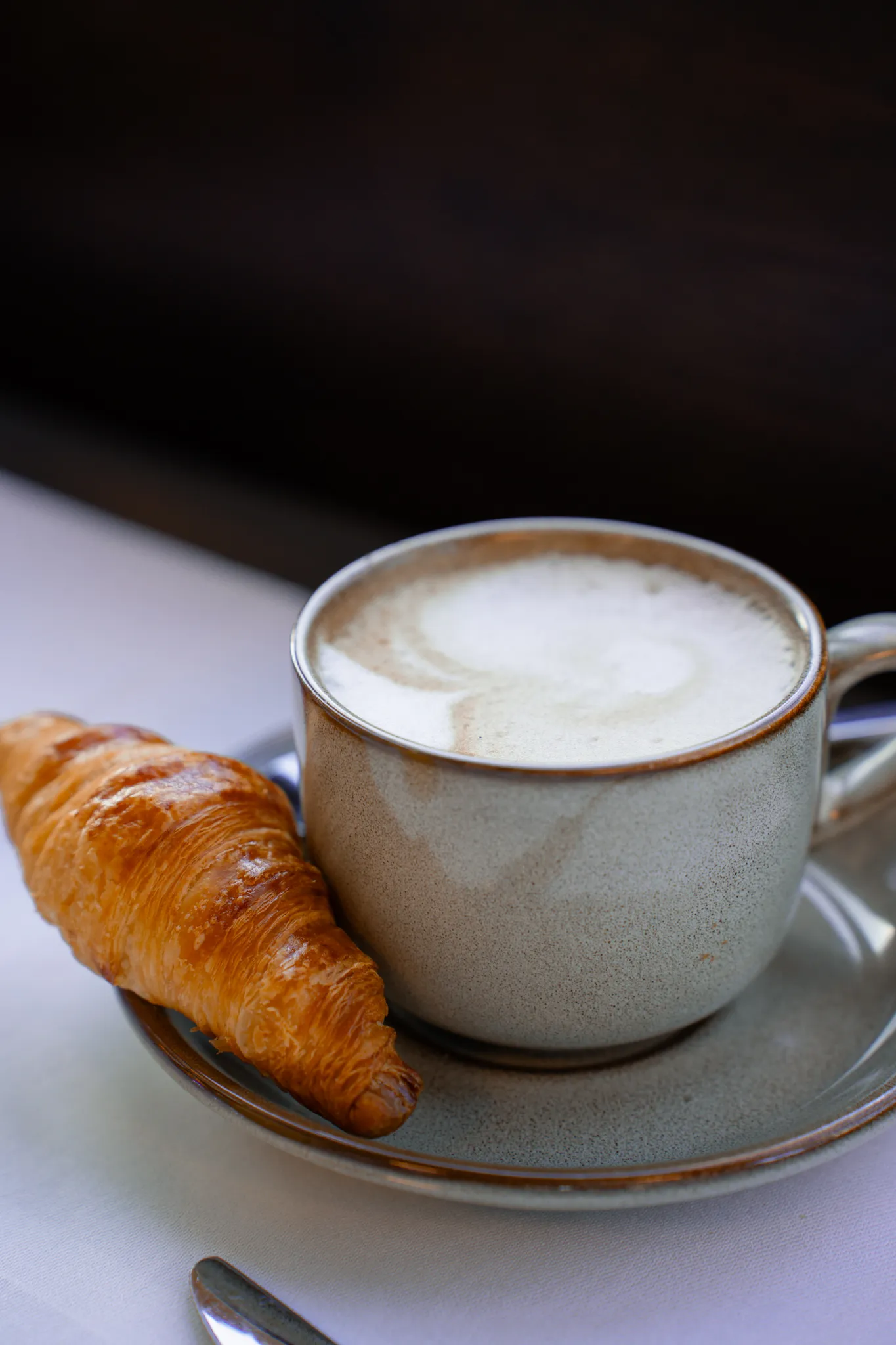 A cup of cappuccino with milk foam and a croissant stand on a grey plate on a table.