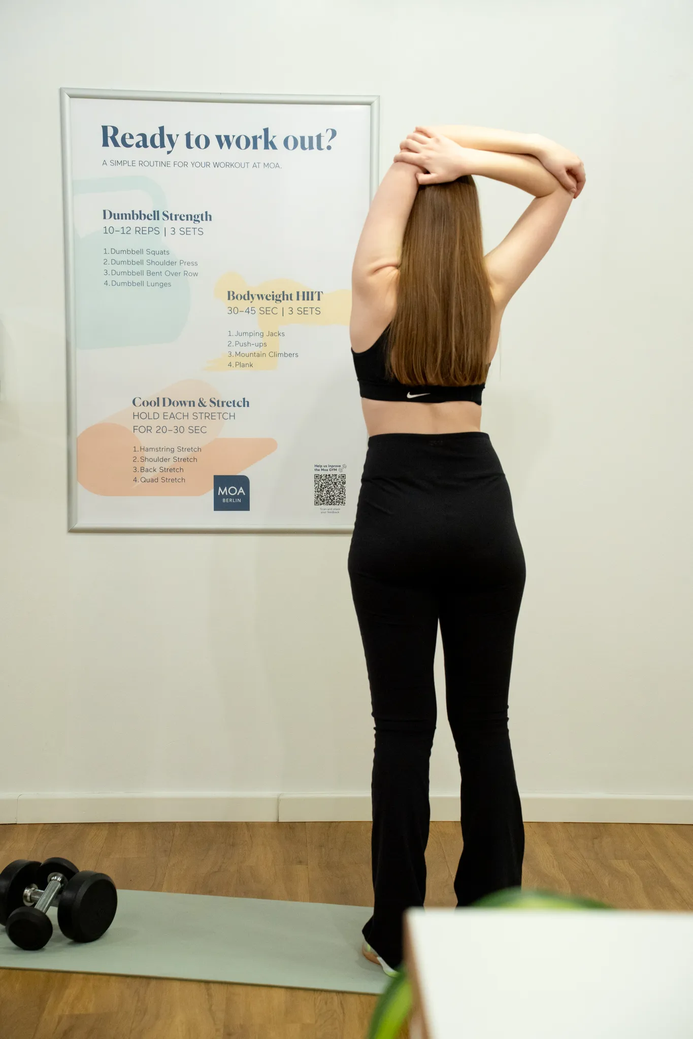 Eine Frau in schwarzer Sportkleidung dehnt sich auf einer Yogamatte vor einem Trainingsplan an der Wand.