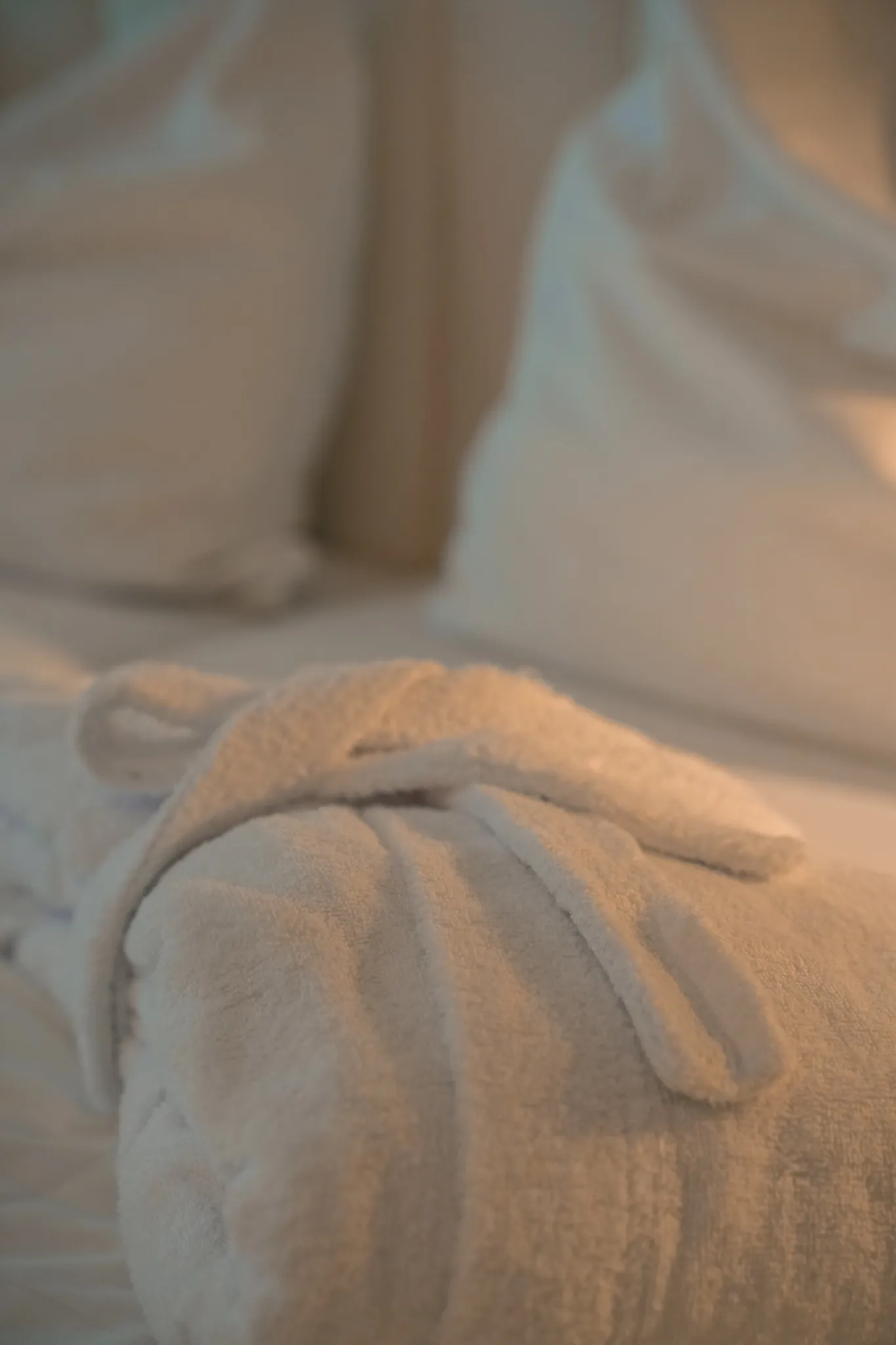 A rolled-up white towel lies on a white-sheeted bed with pillows in warm light.