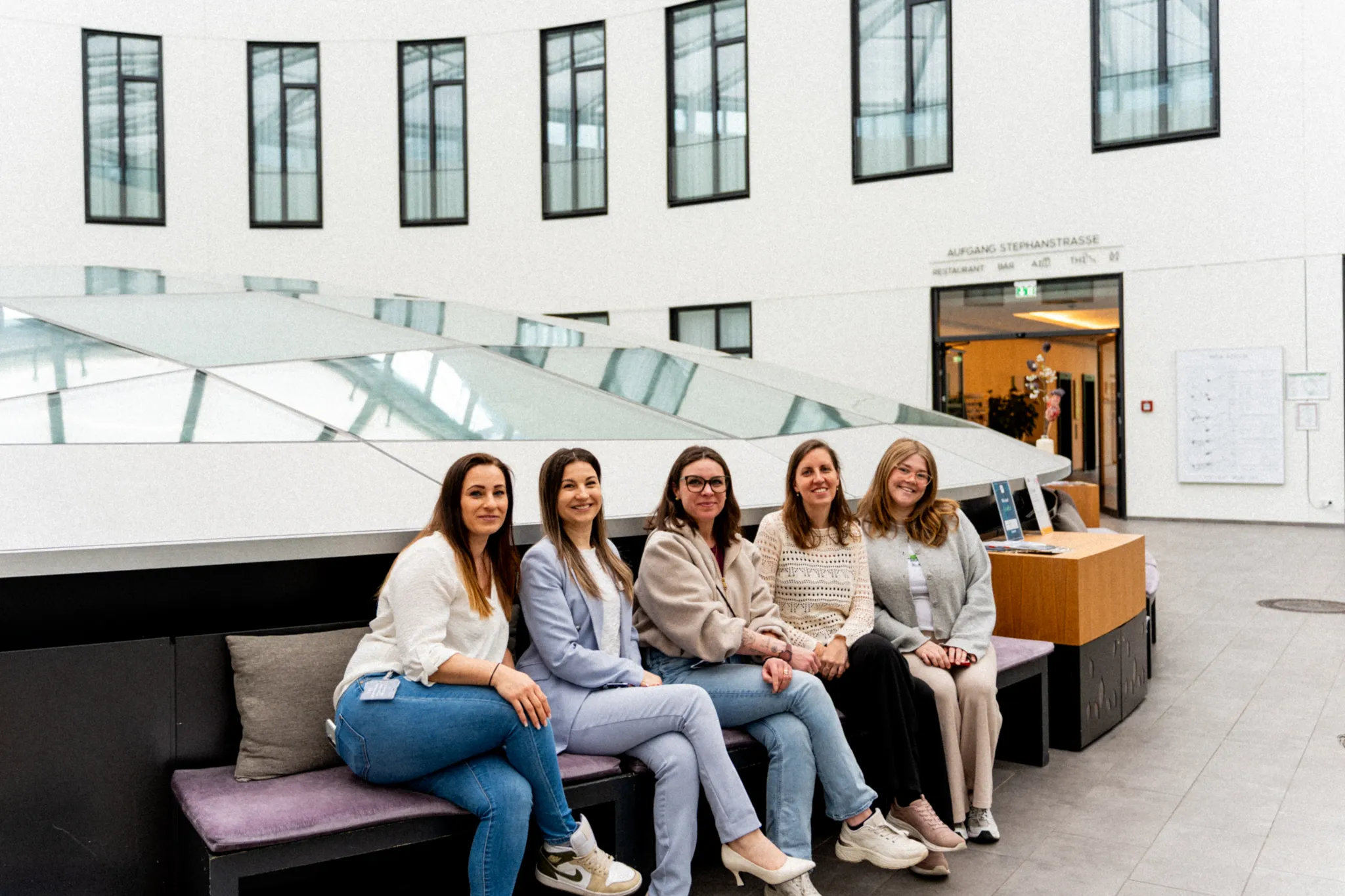 Fünf Frauen sitzen auf einer gepolsterten Sitzbank in der modernen Lobby eines Hotels mit Glasdach und Empfangstresen.