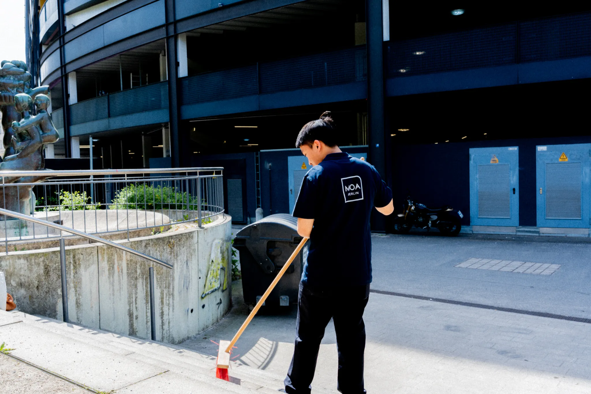 A person sweeps the sidewalk with a broom in front of a modern building, next to a planted concrete basin and parked motor scooters.