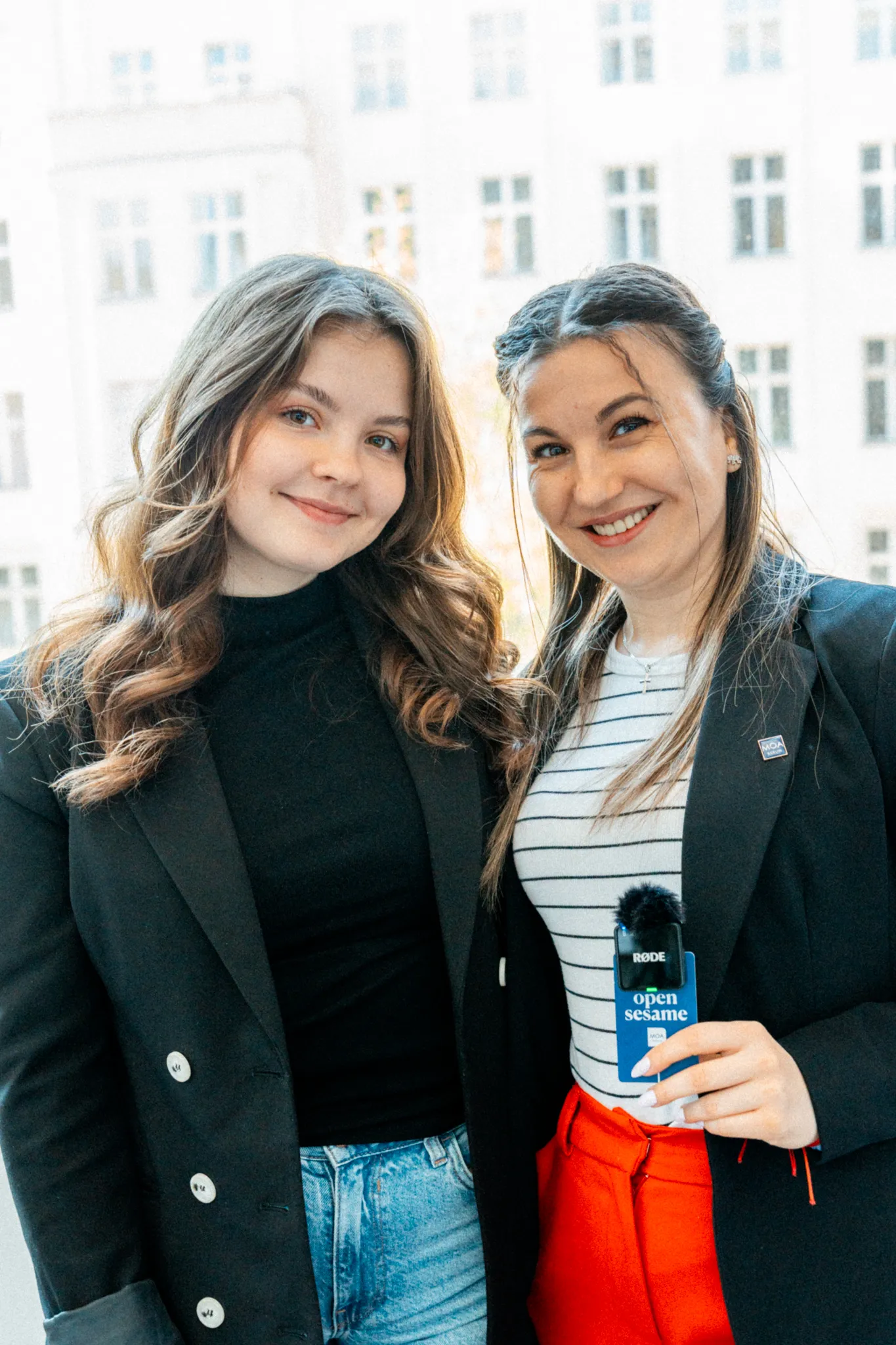 Two women stand in front of a bright building and together hold a microphone with the words "open sesame".
