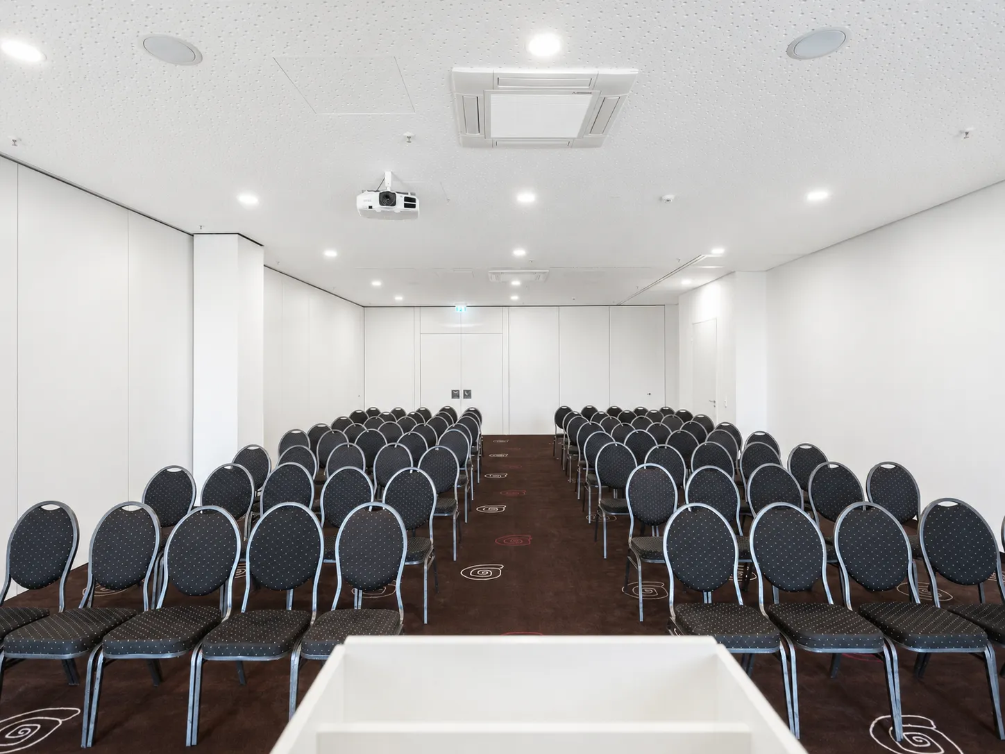 Bright conference room with rows of black chairs, projector, ceiling lights and a white lectern.