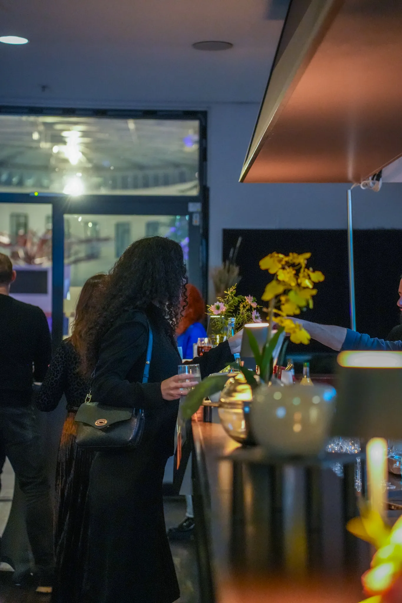 Mehrere Personen stehen an einer Hotelbar mit Blumenarrangements, Getränken und einer beleuchteten Glasfront im Hintergrund.