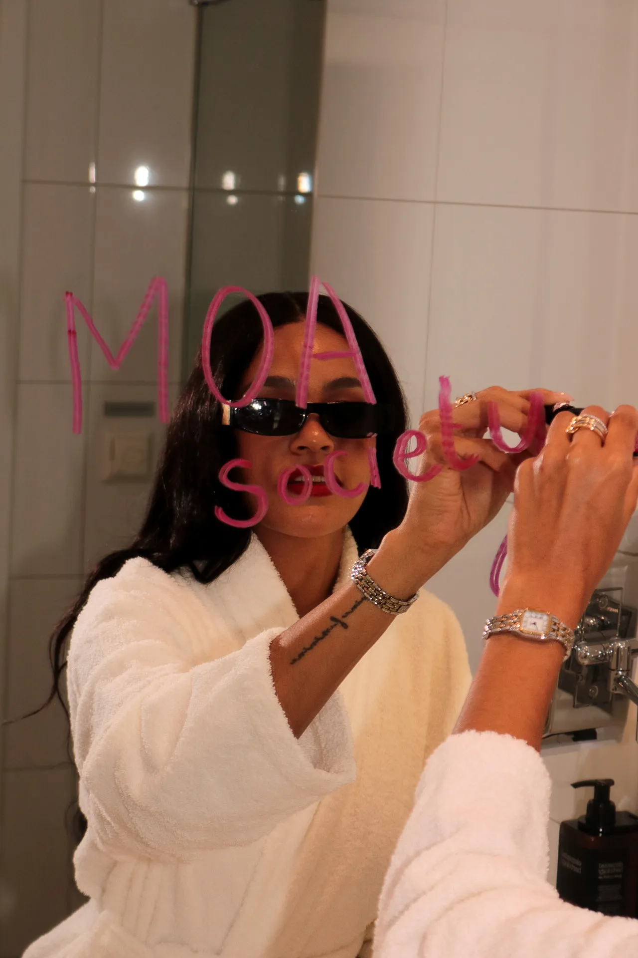 A person in a white bathrobe writes "MOA" and "Souls" with lipstick on a bathroom mirror.