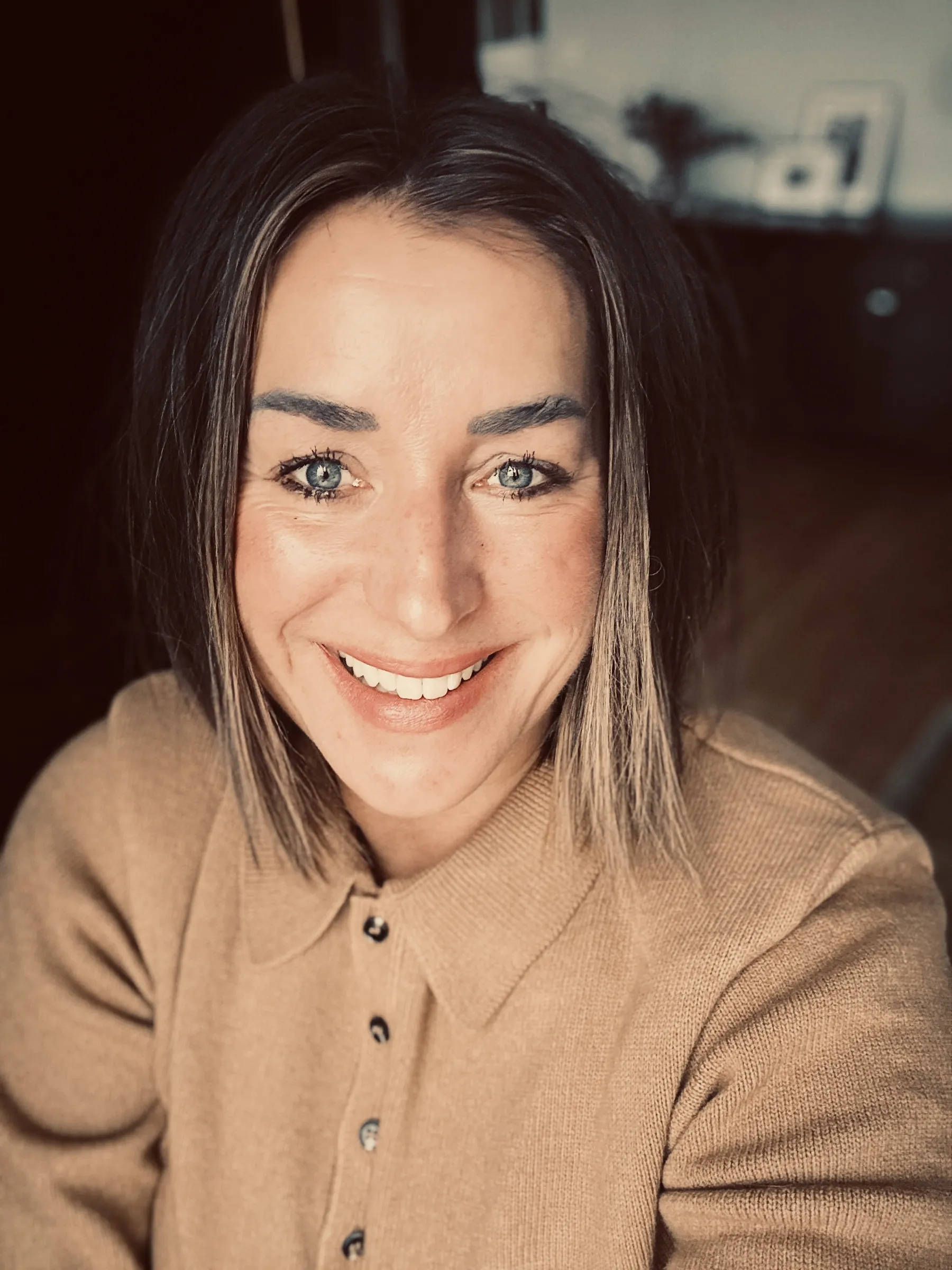 A smiling woman with shoulder-length brown hair wears a beige sweater with a collar and looks at the camera.