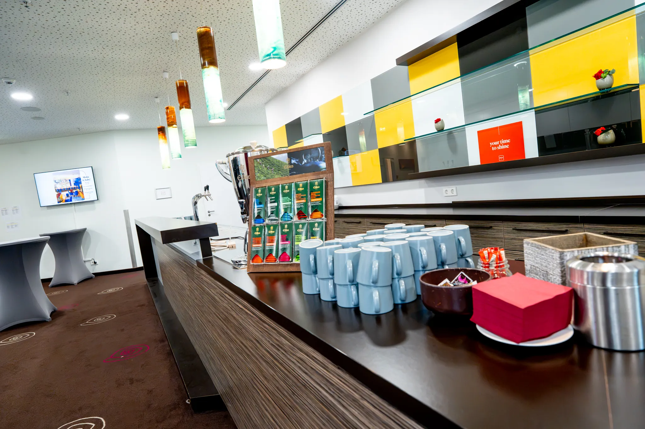 Moderne Hotelbar mit Theke, gestapelten blauen Tassen, Teebeutelspender und bunten Wandpaneelen.