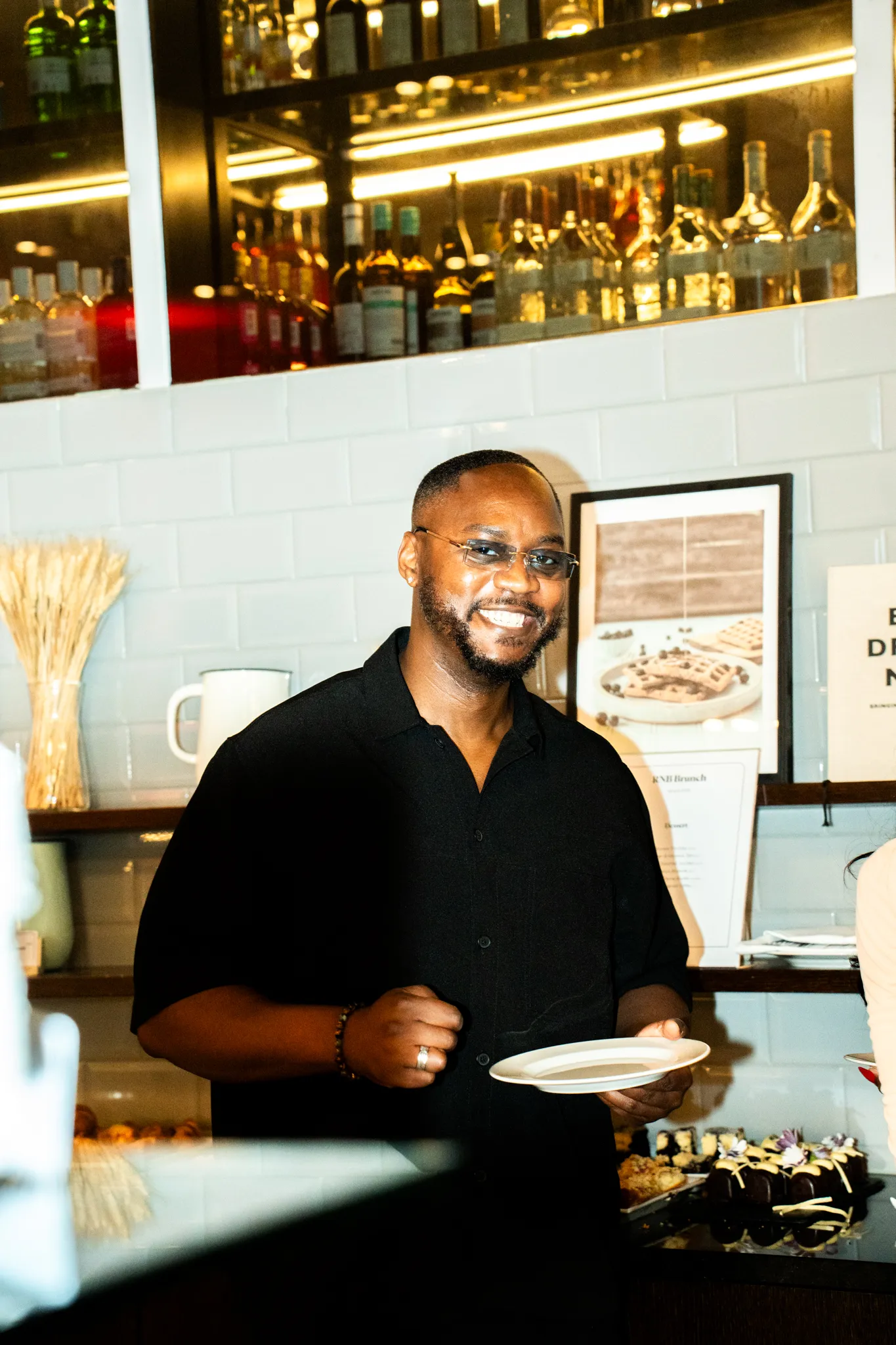 A man in a black shirt holding a plate in a café with drink bottles and food in the background.