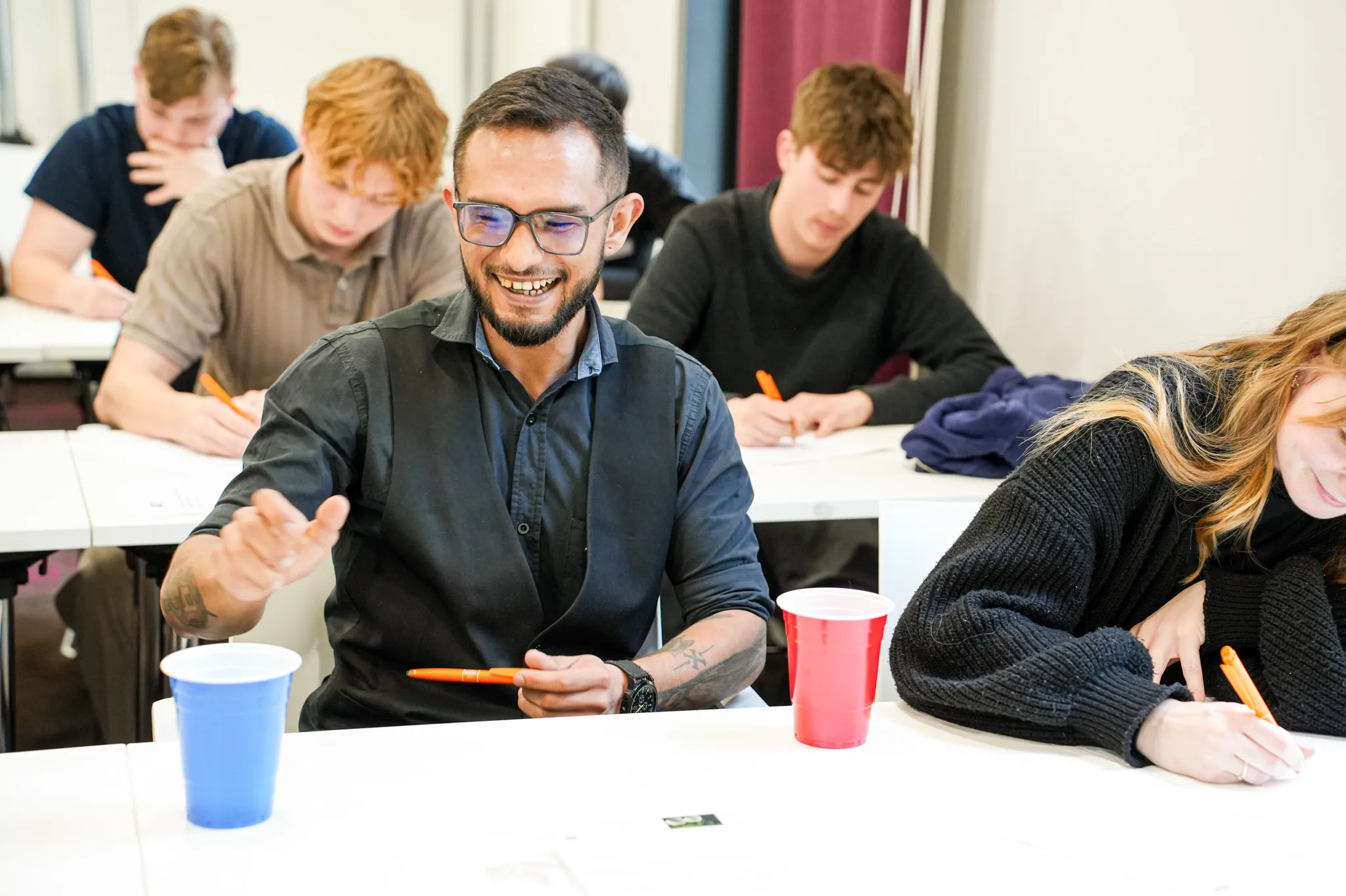 Several young people sit at tables and write, while a man in the foreground holds an orange pen.