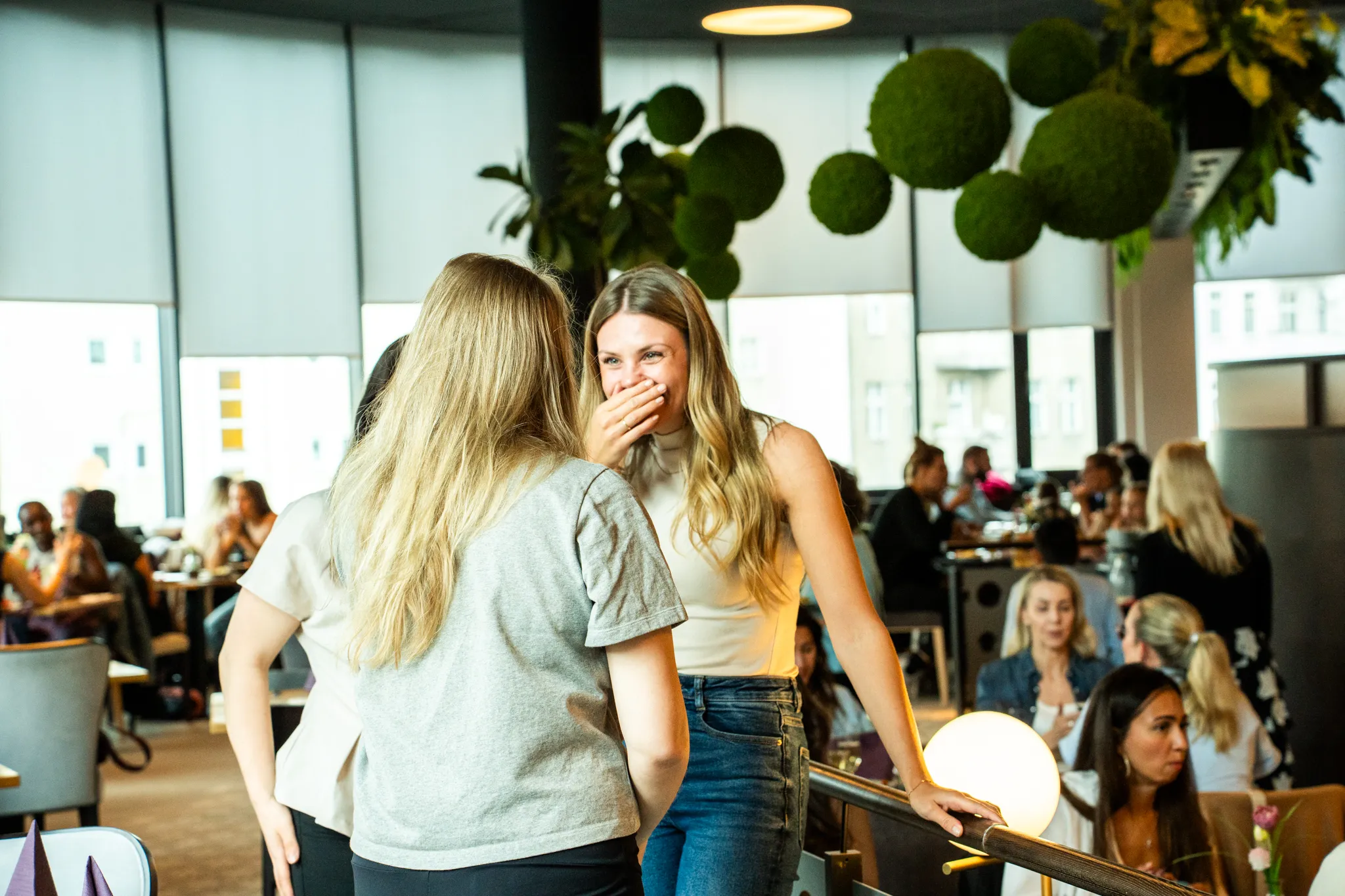 Zwei Frauen stehen in einem hellen Restaurantbereich mit Gästen, Tischen, Pflanzen und großen Fenstern.