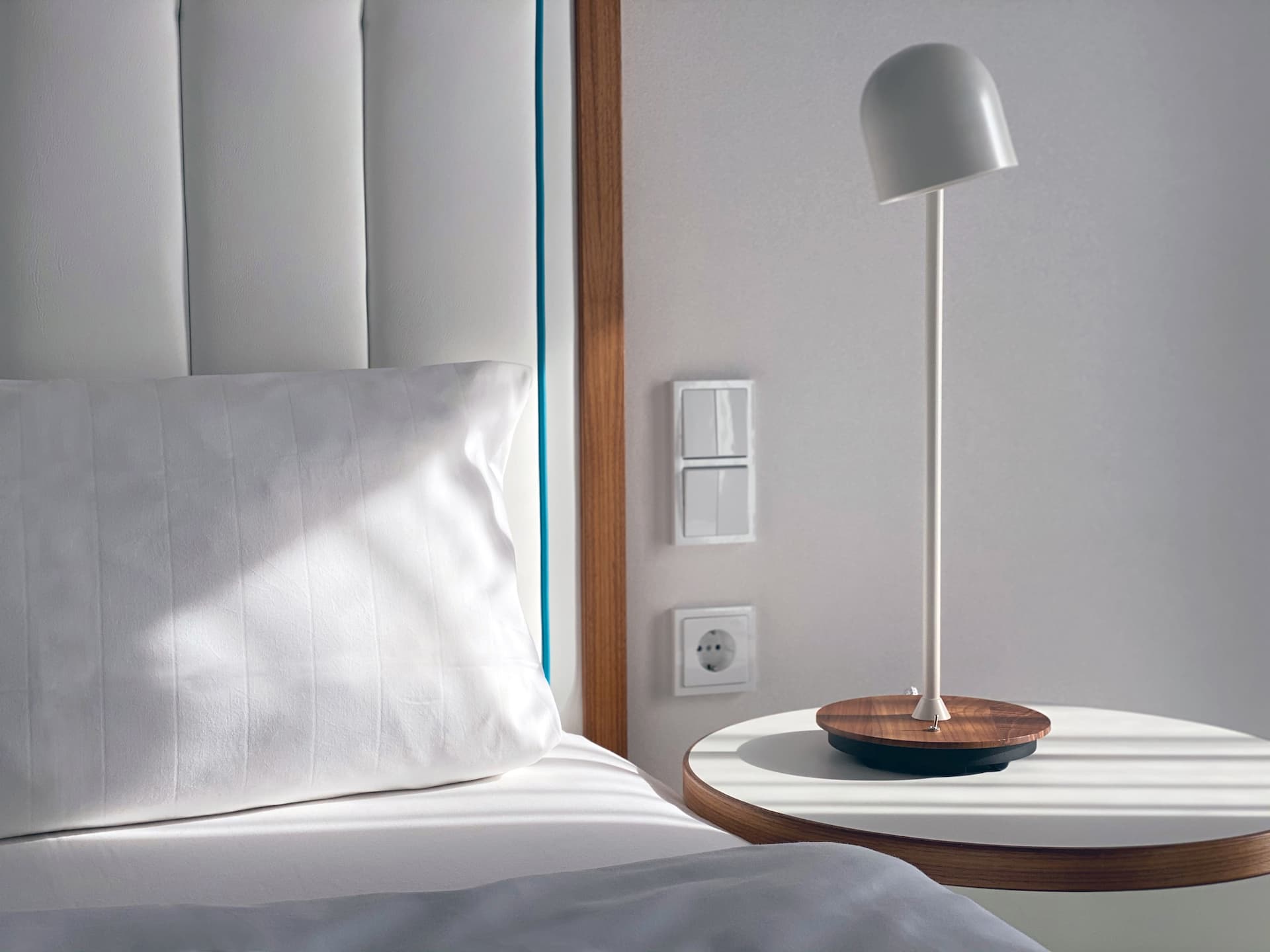 A white hotel bed with pillows next to a round side table, on which stands a modern table lamp in front of a light-coloured wall.