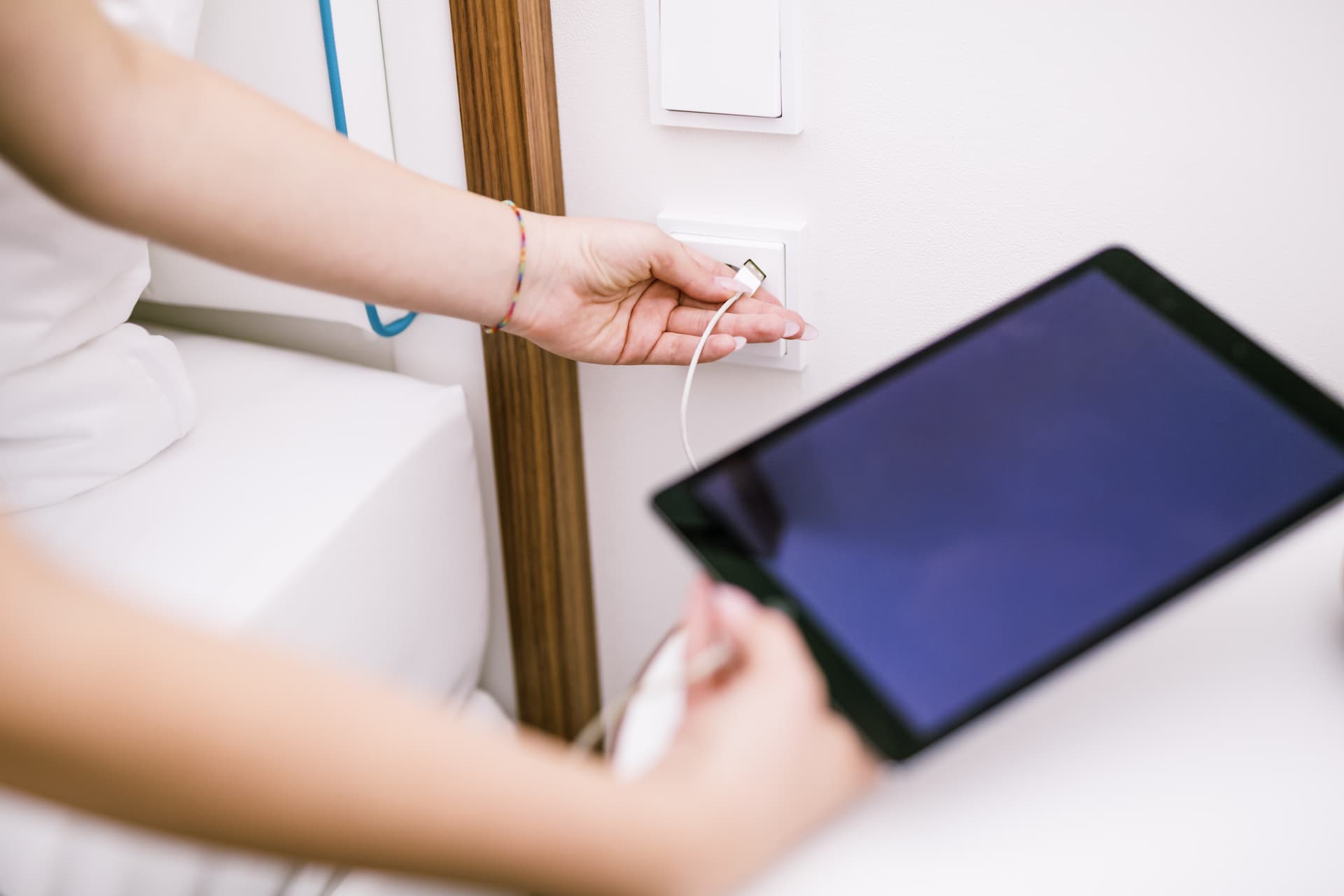 Eine Person hält ein Tablet, während ein Ladekabel an einer Steckdose neben einem Bett angeschlossen wird.