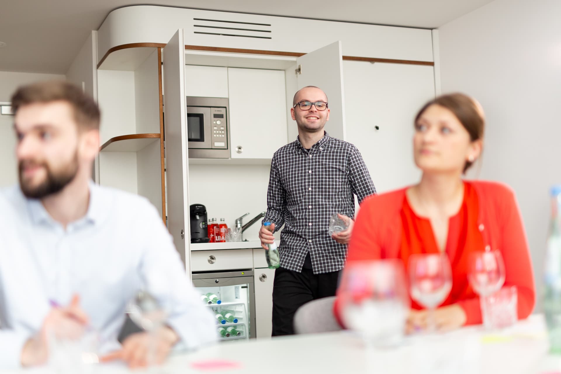 Drei Personen sitzen und stehen in einer Hotelküche mit Einbaugeräten, Getränken und Gläsern auf einem Tisch im Vordergrund.