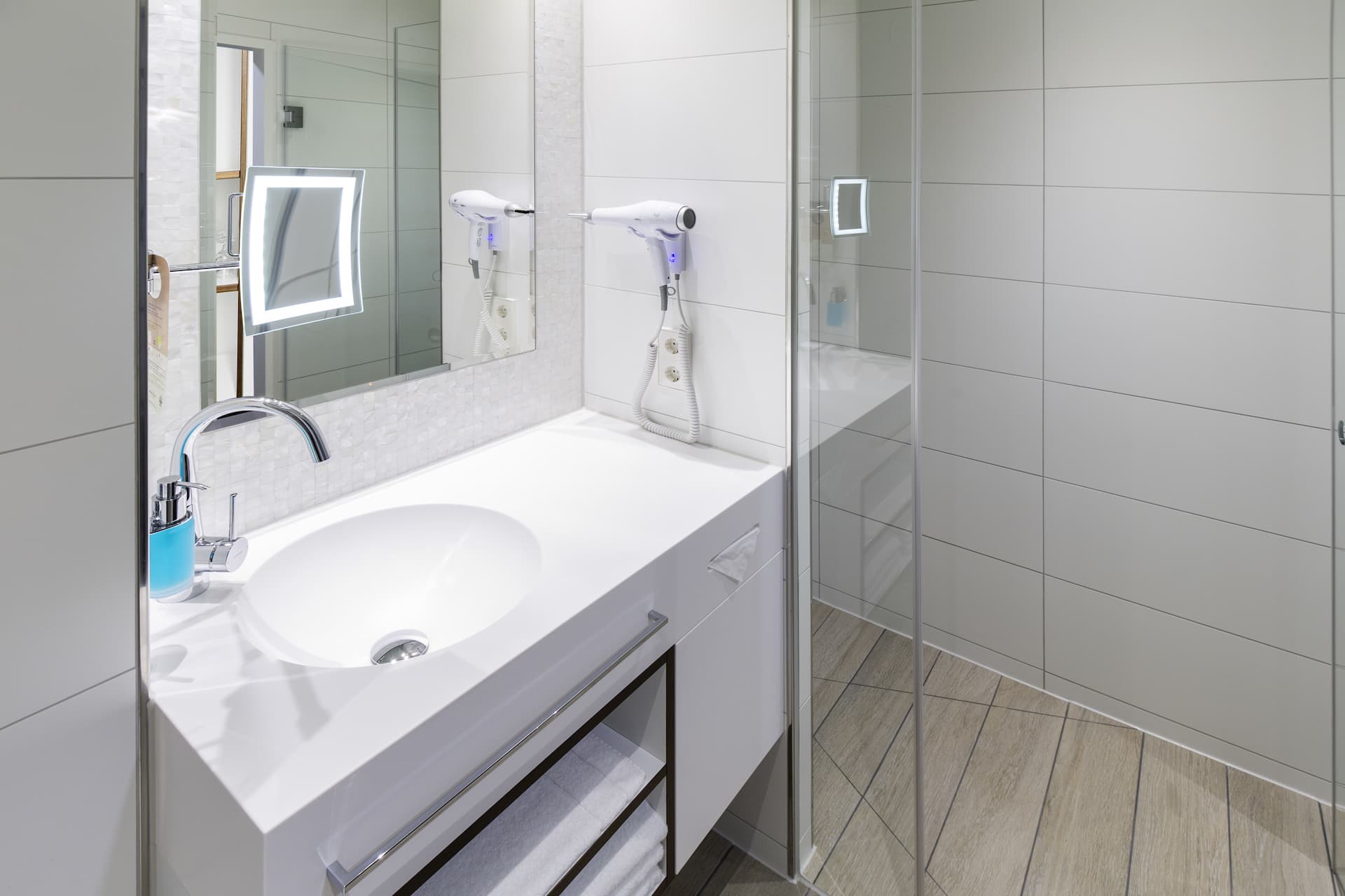 Modernes Badezimmer mit weißem Waschbecken, großem Spiegel, Wandhaartrocknern, Dusche und hellem Fliesenboden.