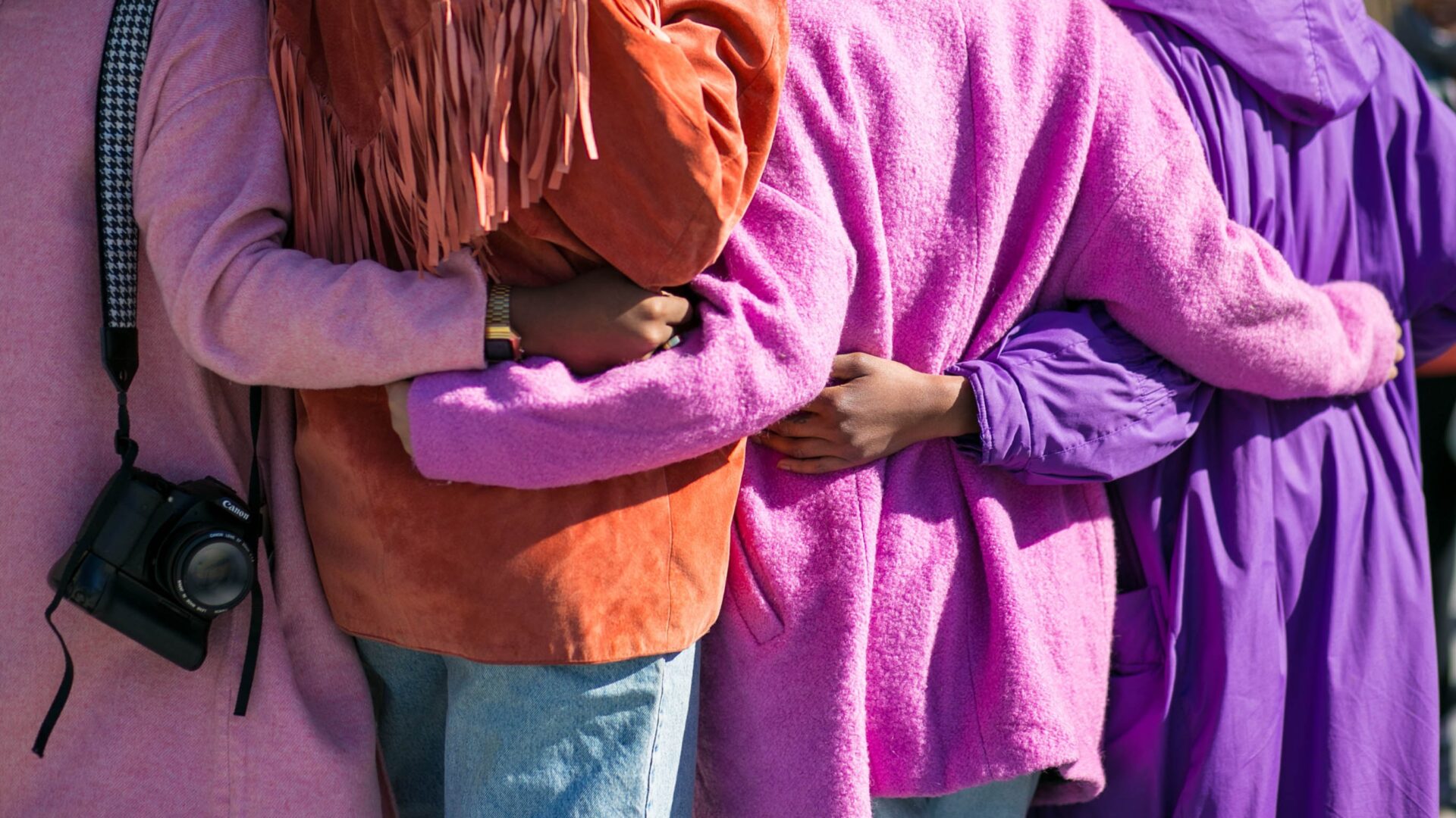 Three people in colourful clothing stand close together; on the left, one person carries a camera over their shoulder.