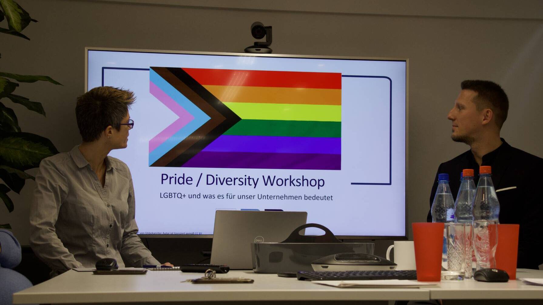 A screen shows a rainbow flag and the text “Pride / Diversity Workshop,” while two people sit at a table.