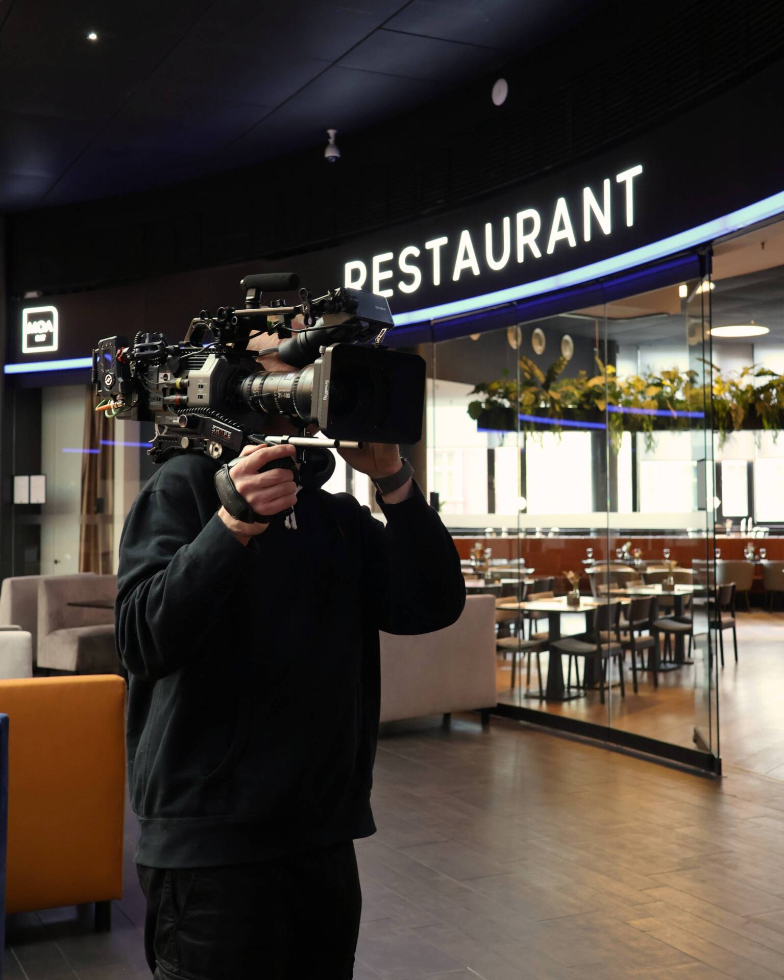 A person films a hotel's restaurant area with a professional camera, with tables, chairs and plants visible.