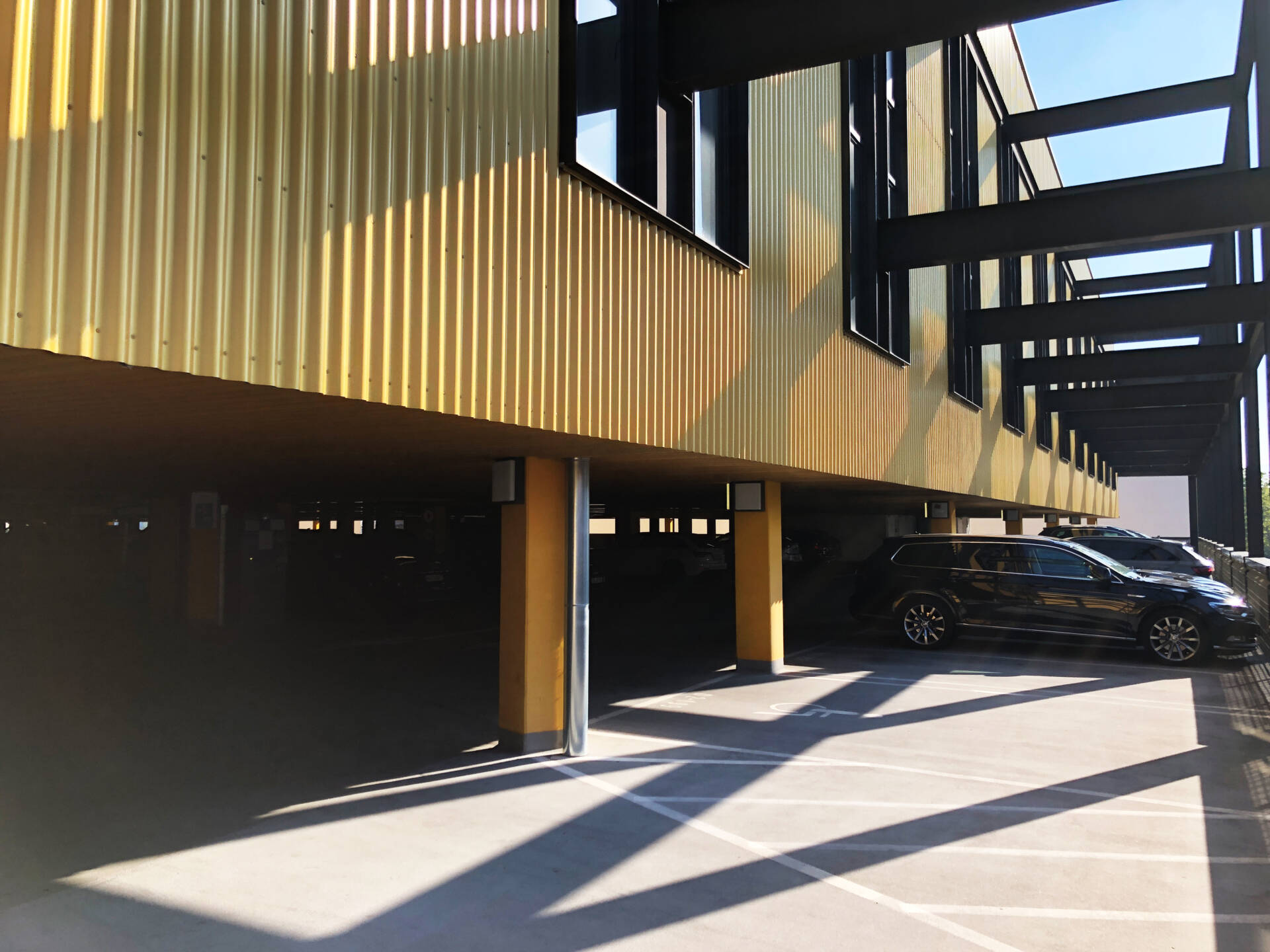 Covered parking area with yellow facade cladding, black steel beams, and a black car in sunlight.