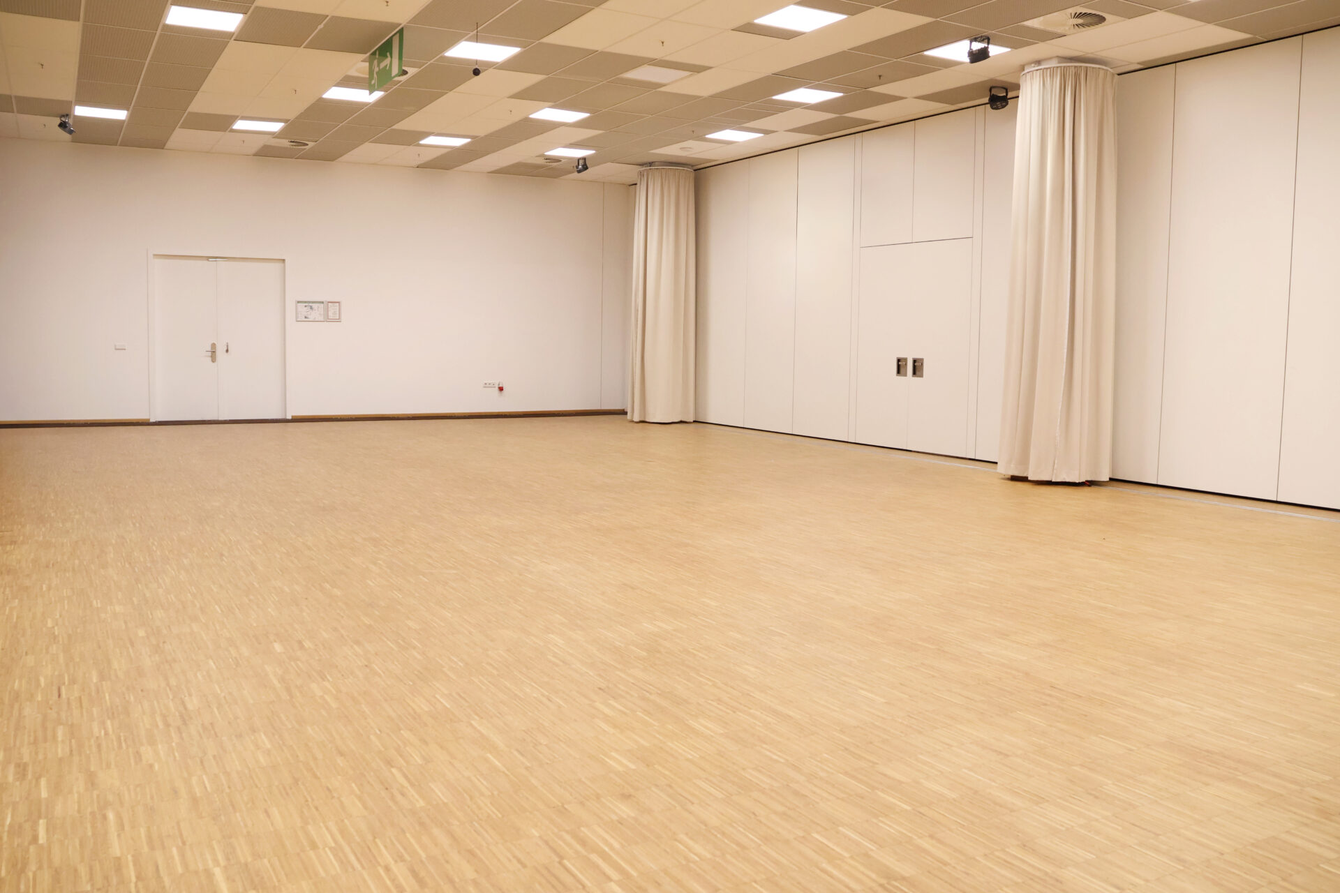 Ein großer, leerer Veranstaltungsraum mit einer hellen Deckenbeleuchtung, Säulen, einem Holzfußboden und flexiblen Trennwänden.