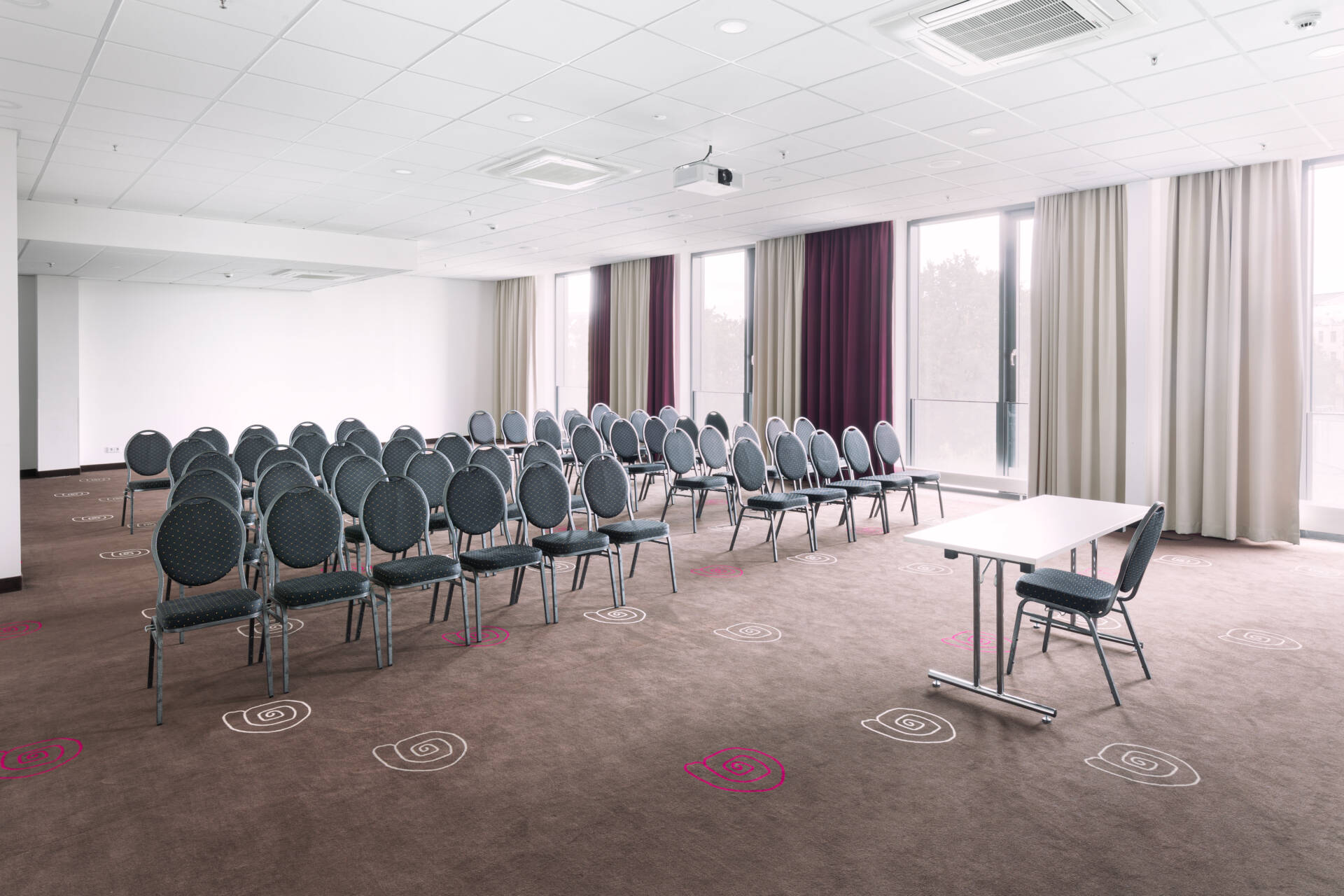 A bright conference room with numerous rows of chairs, individual tables, carpet flooring, curtains and daylight through large windows.