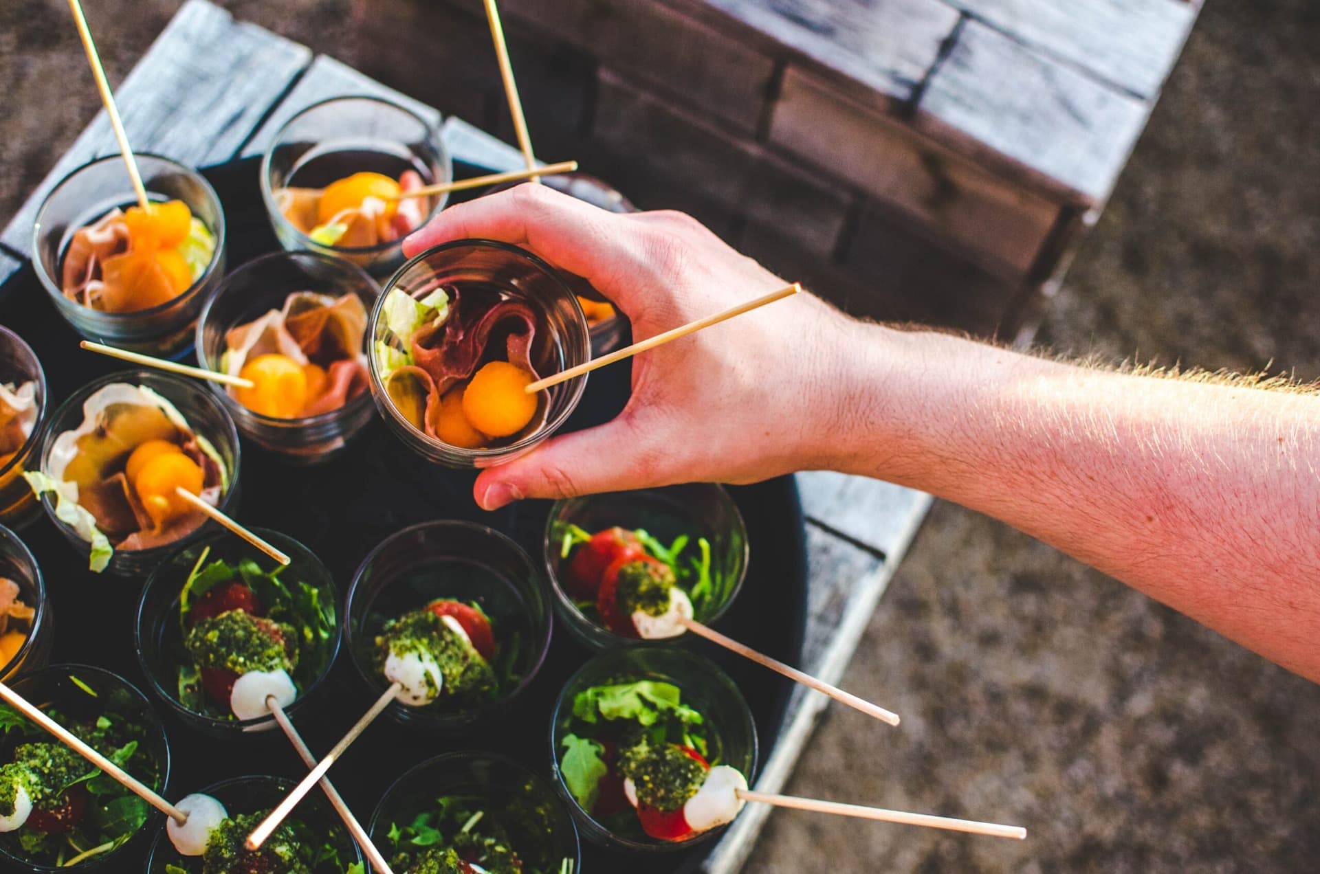 A hand reaching for a glass with small skewers of vegetables and fruit on a round tray at an event venue in Berlin Mitte.