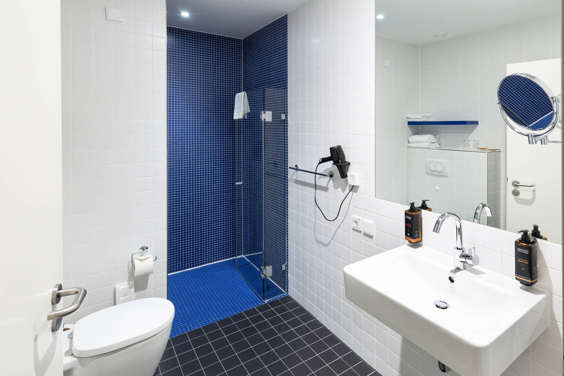Helles Badezimmer mit weißem Waschbecken, bodengleicher Dusche, Toilette, großem Spiegel und blau-weißen Fliesen.