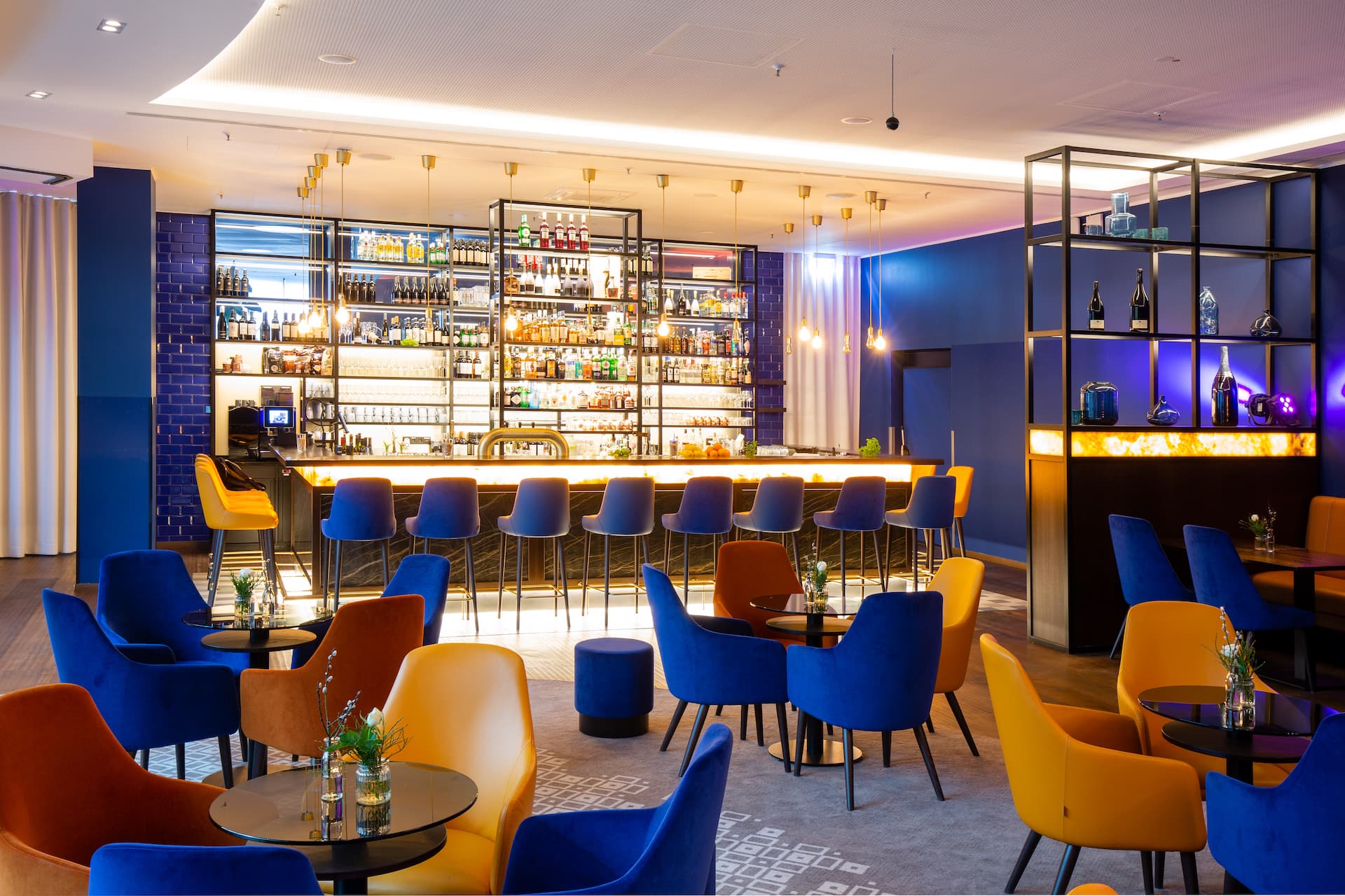 A hotel bar in Berlin with tall bar stools in blue and yellow, several tables with chairs as well as decorative shelves and indirect lighting.