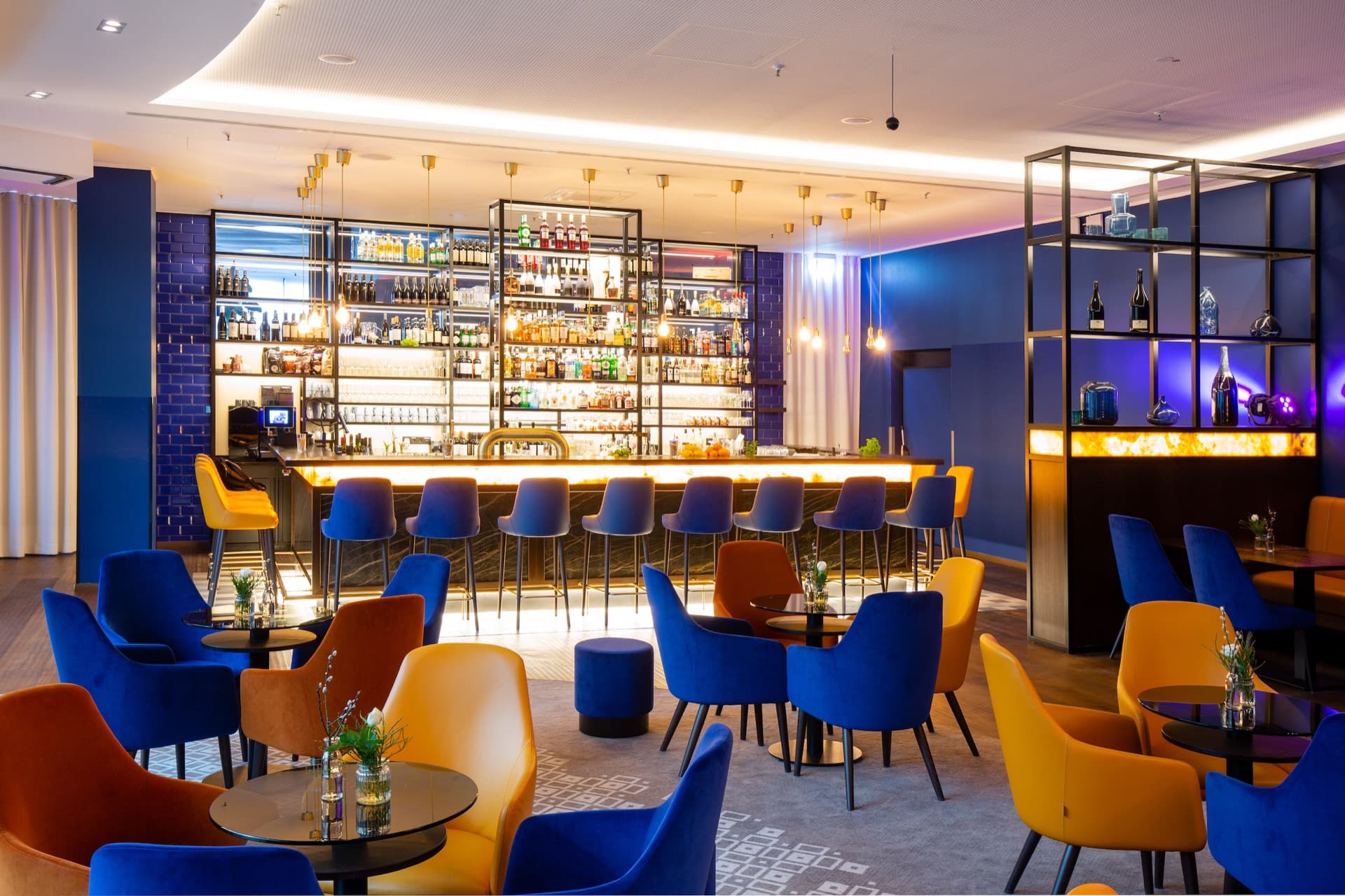 A hotel bar in Berlin with tall bar stools in blue and yellow, several tables with chairs as well as decorative shelves and indirect lighting.