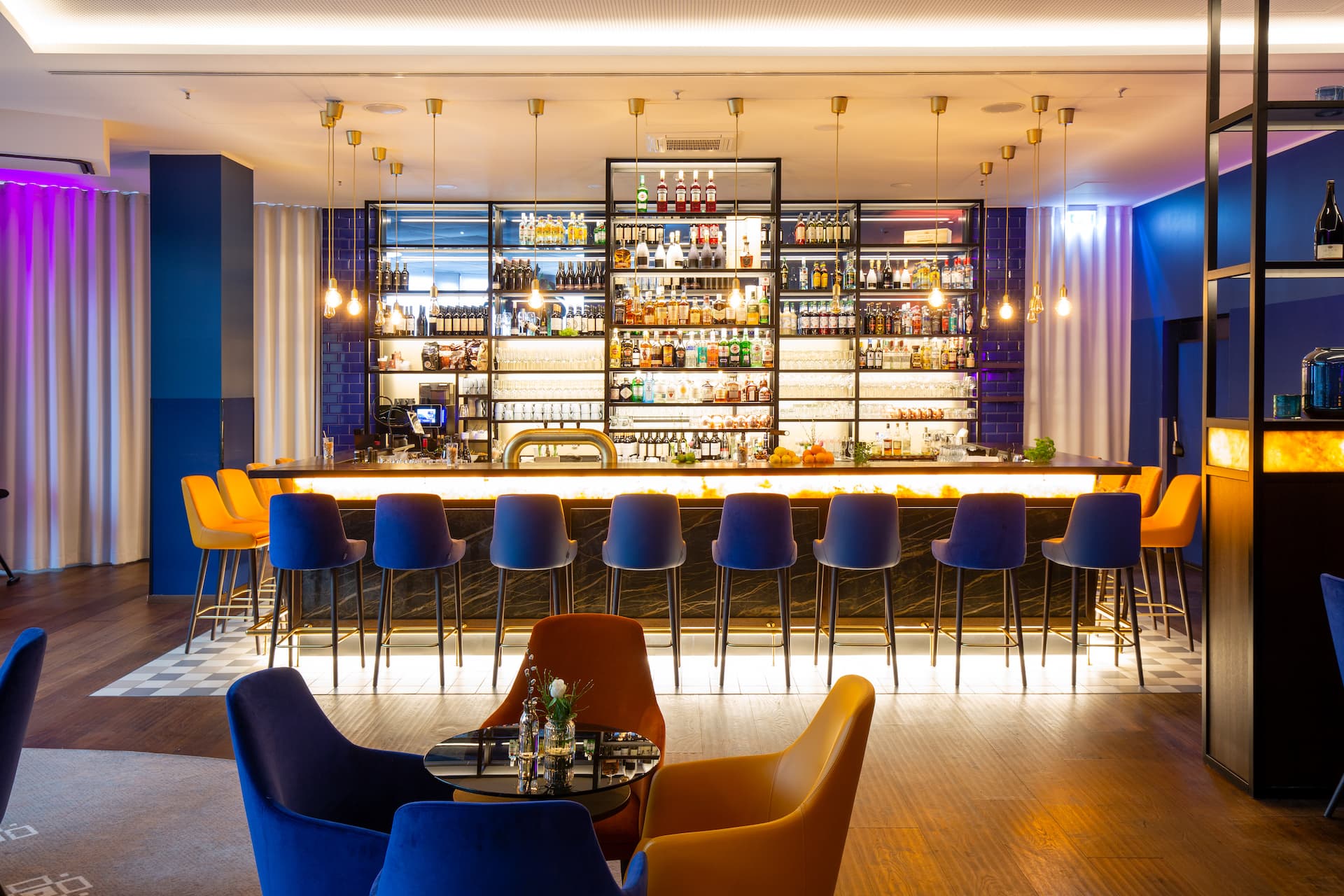 A hotel bar in Berlin with a long counter, yellow and black bar stools and shelves of bottles in the background, with seating areas featuring blue and yellow armchairs in front.