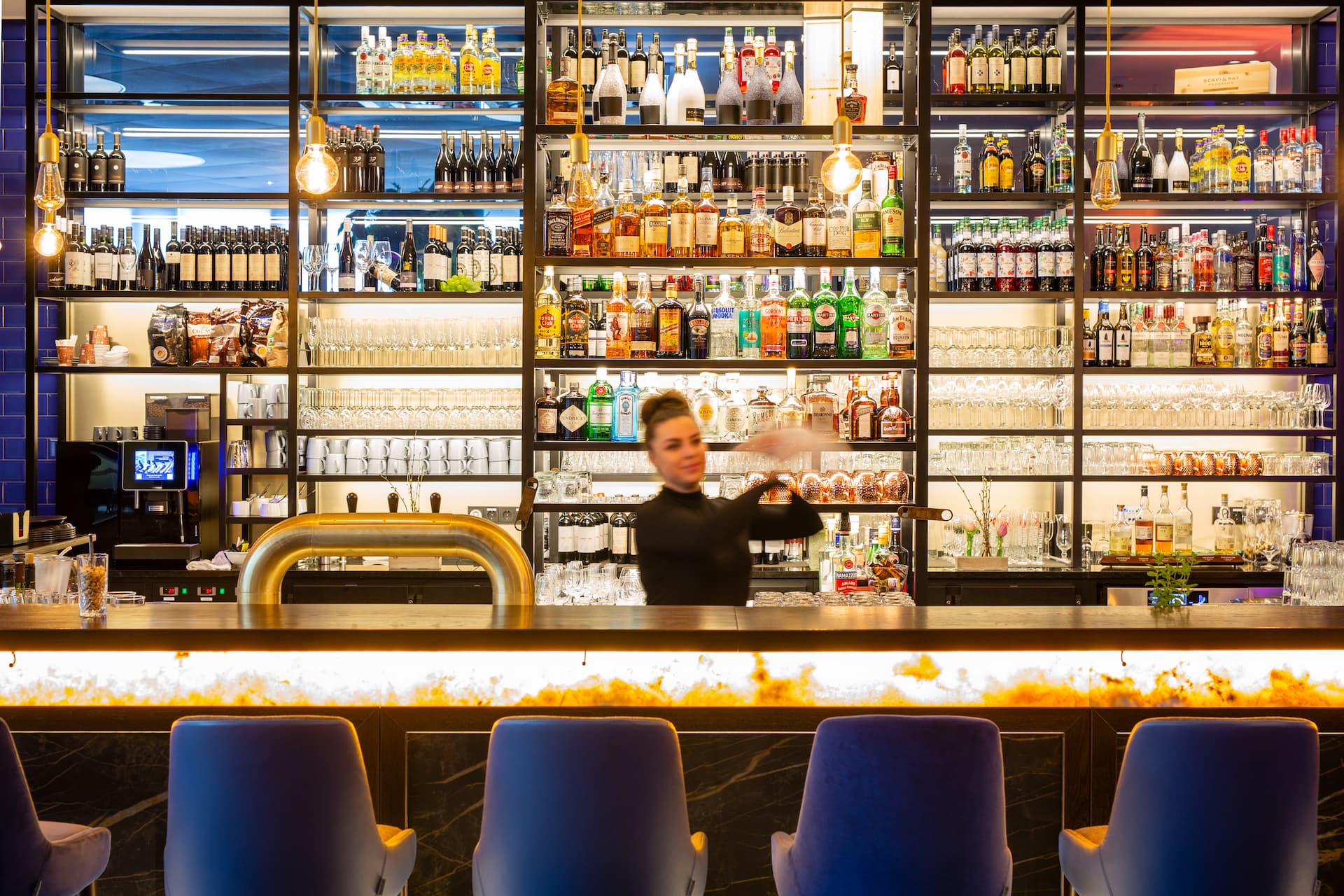 Eine Barkeeperin mischt einen Cocktail hinter einer Bar mit beleuchtetem Tresen und Regalen voller Spirituosen und Gläser.
