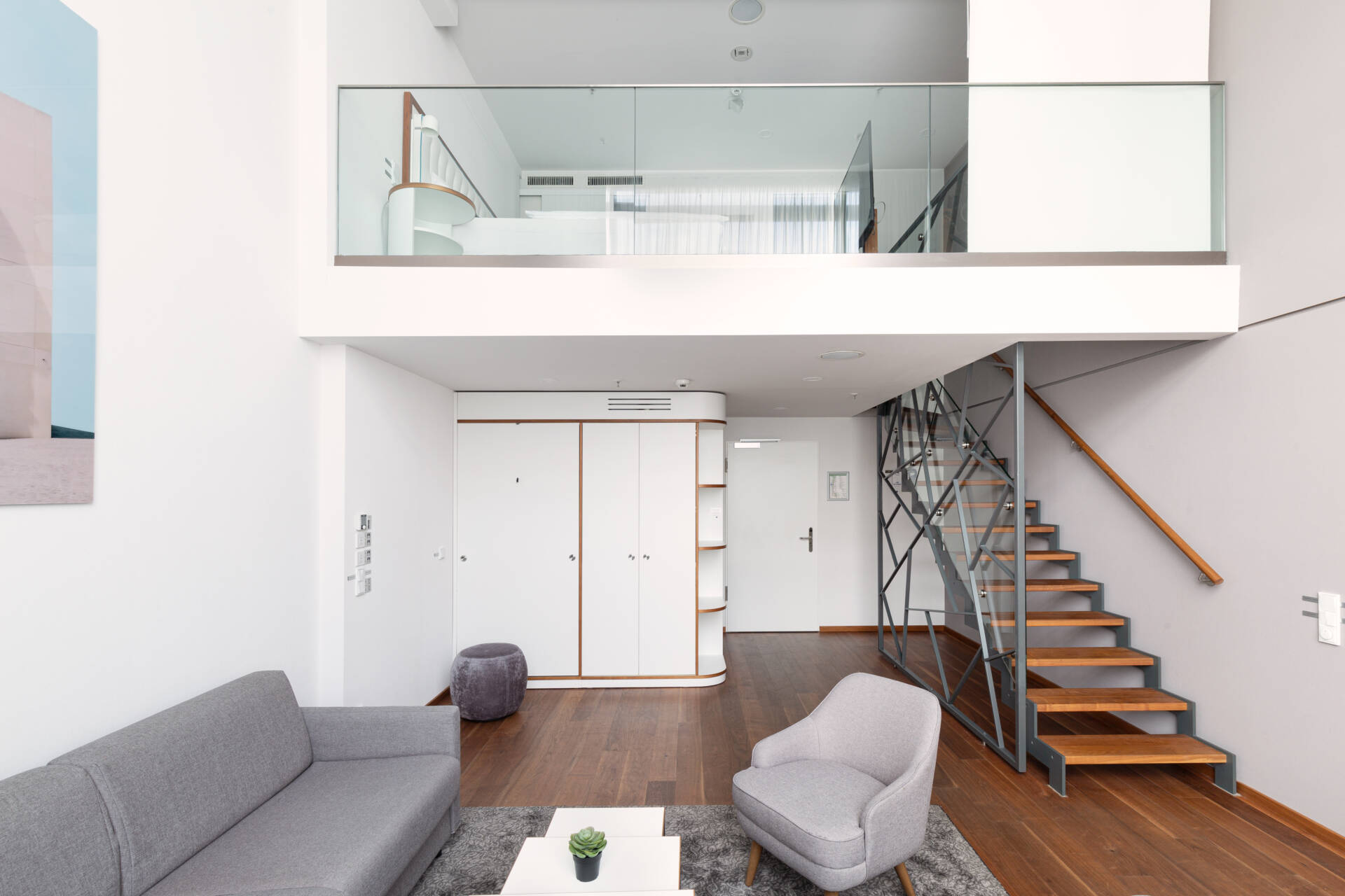 Modern hotel suite with grey sofa, armchair, kitchenette, wooden staircase and glazed gallery.