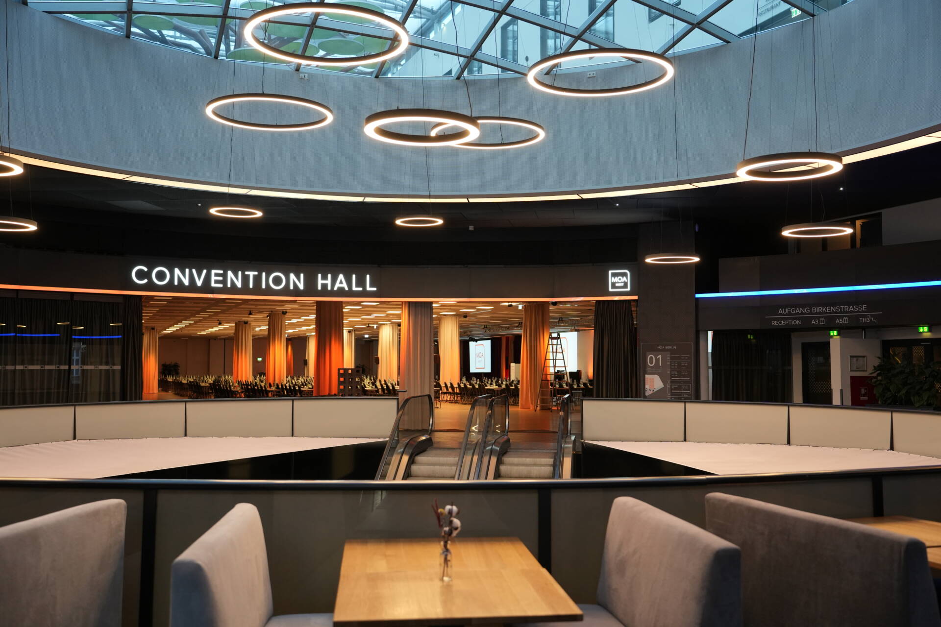 A modern convention hall with escalators, seating areas, round ceiling lighting, and a view into a prepared event space.