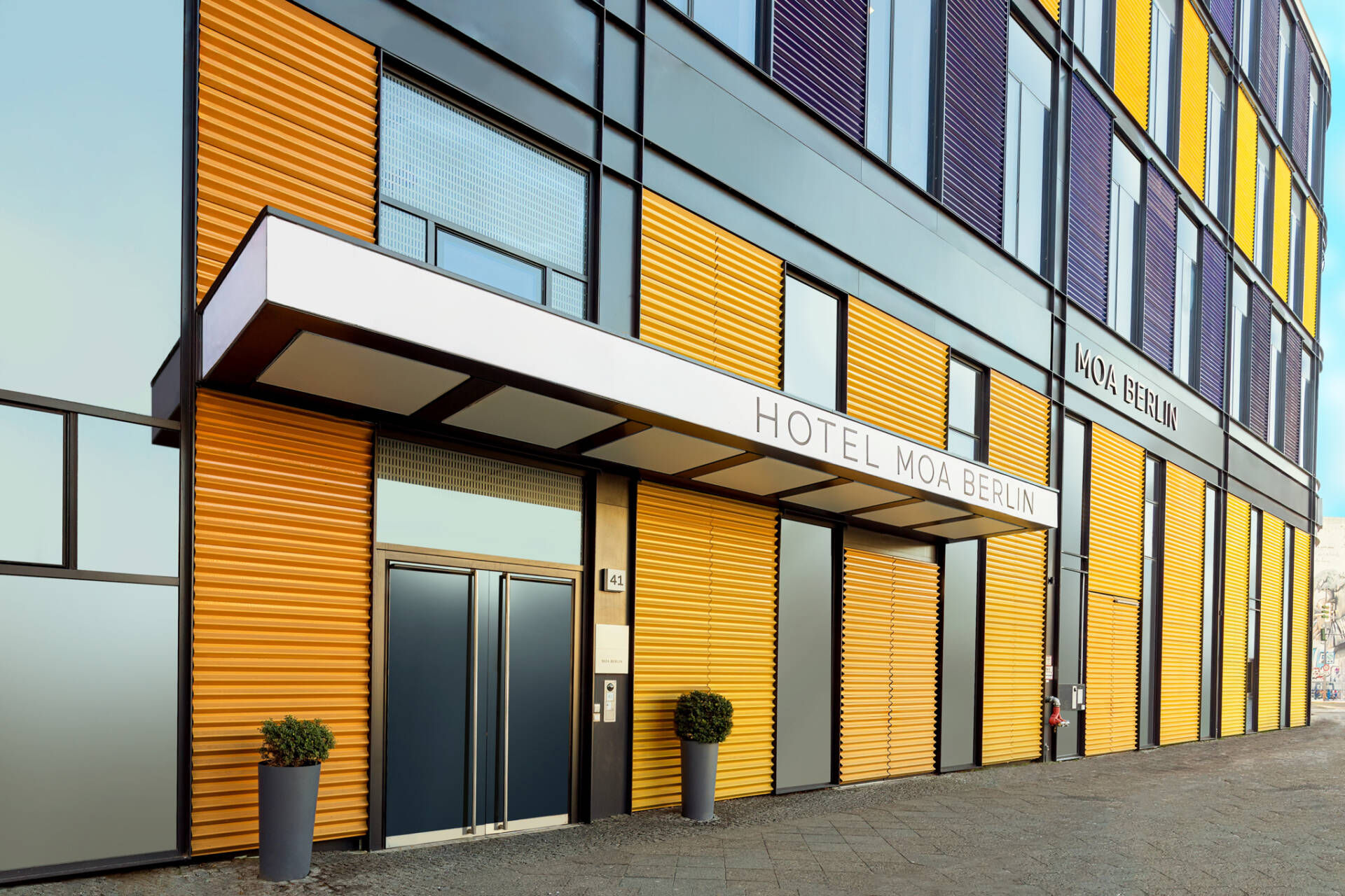 Der Eingangsbereich des Hotels MOA Berlin mit gelber Fassade, Vordach, Türen, Pflanzkübeln und dem Schriftzug „HOTEL MOA BERLIN“.