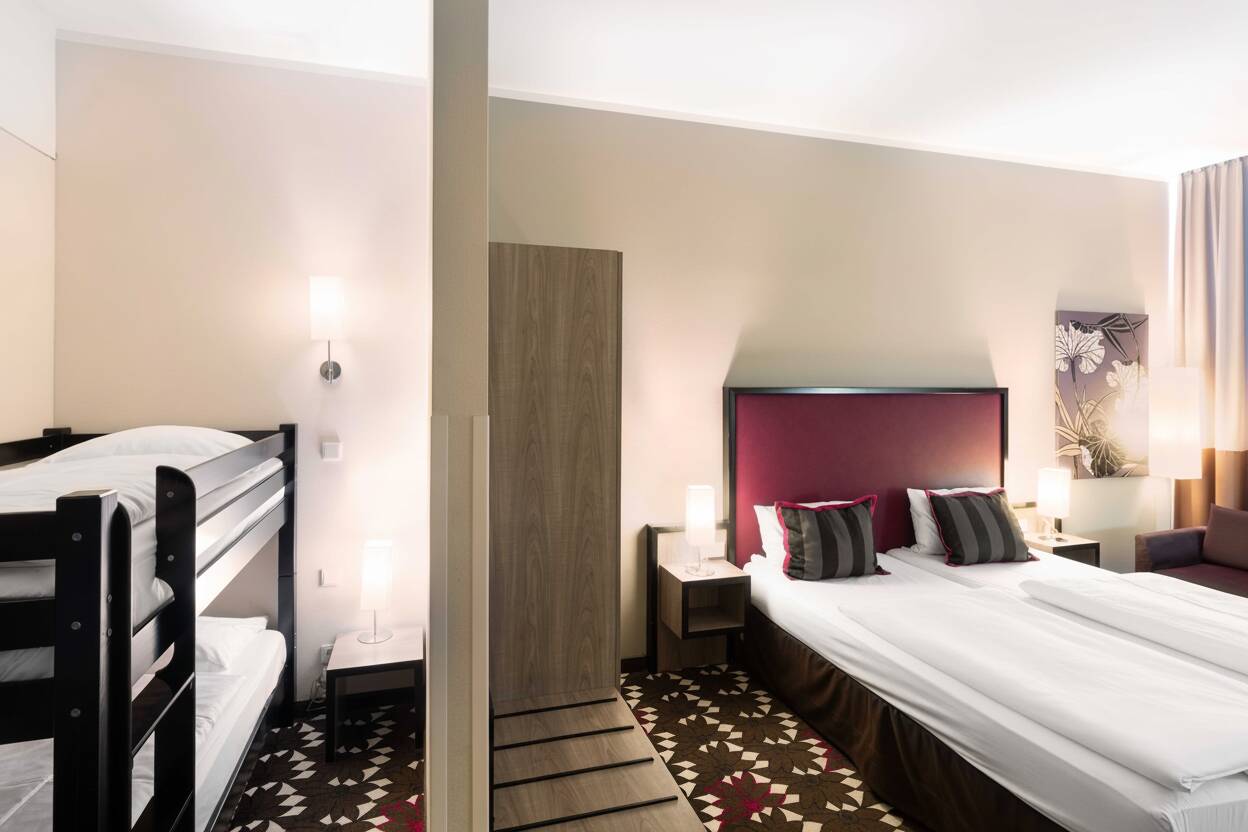 Modernes Hotelzimmer mit Doppelbett, Hochbett, Nachttischen, Kleiderschrank und gemustertem Teppichboden.