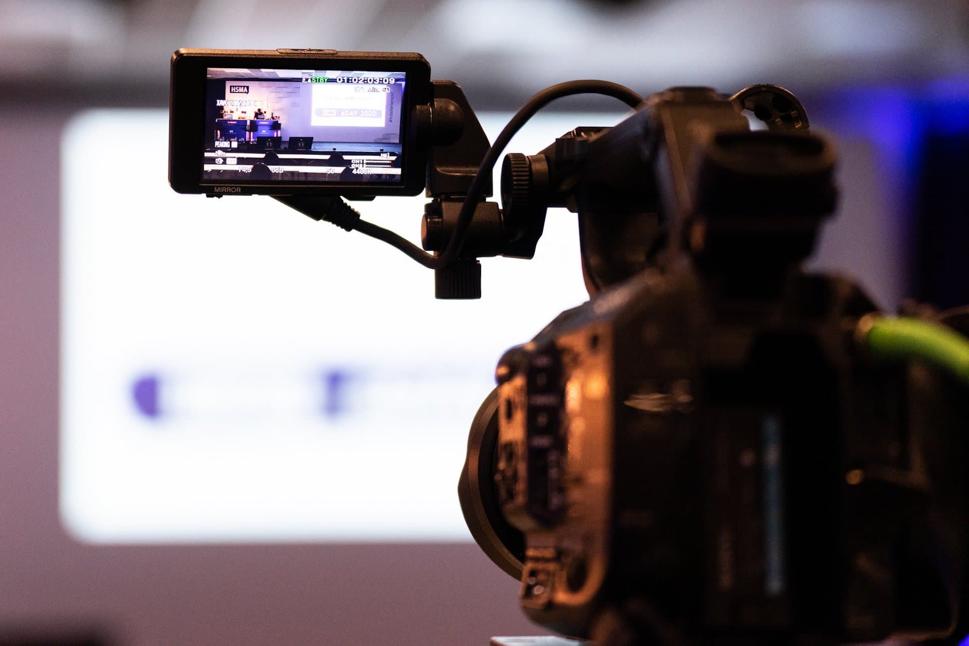 Eine professionelle Videokamera filmt eine Präsentation oder Veranstaltung in einem Konferenz- oder Eventraum mit unscharfem Hintergrund.