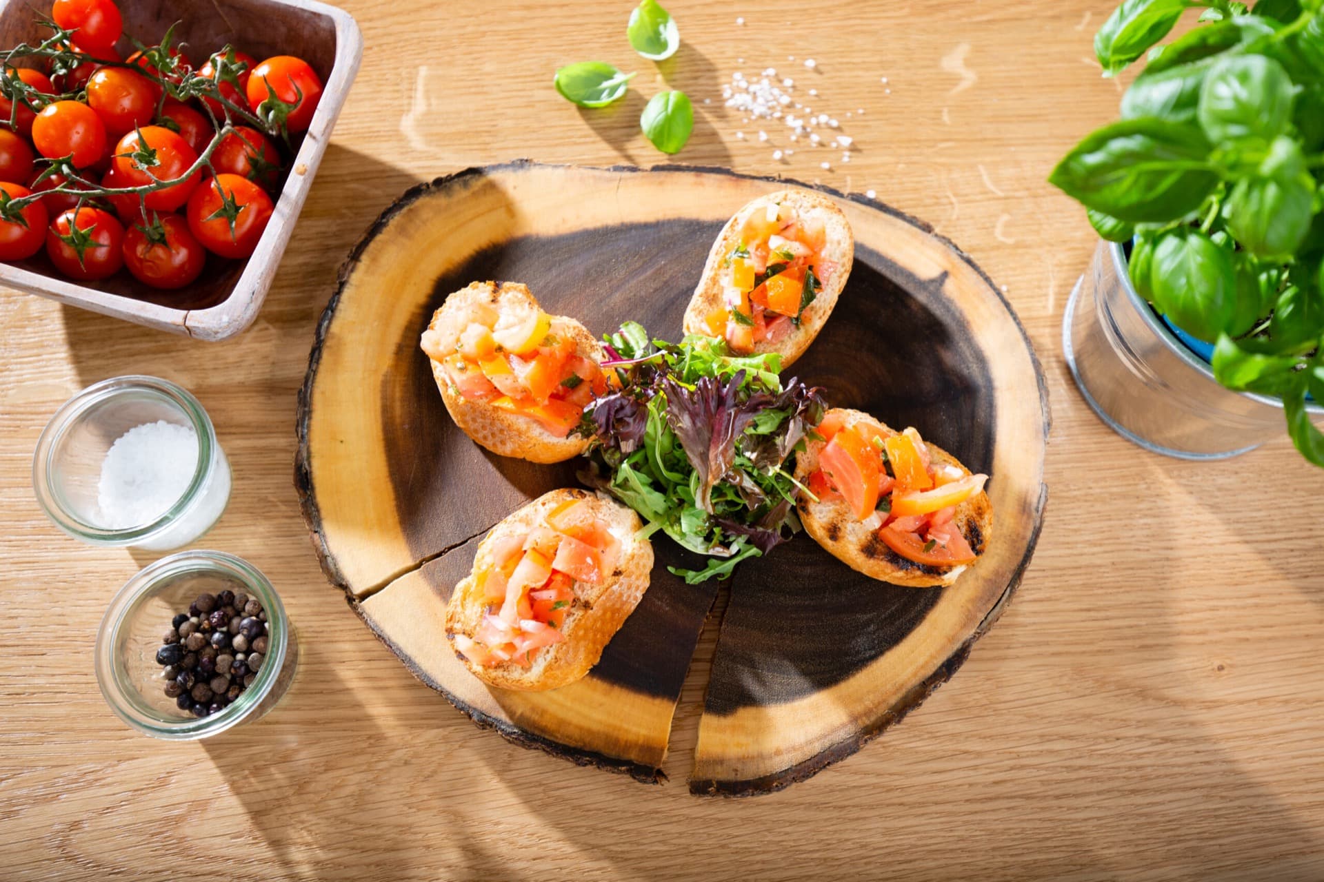 Four bruschetta with tomatoes and herbs on a rustic wooden board, alongside fresh tomatoes, pepper, salt, and basil.