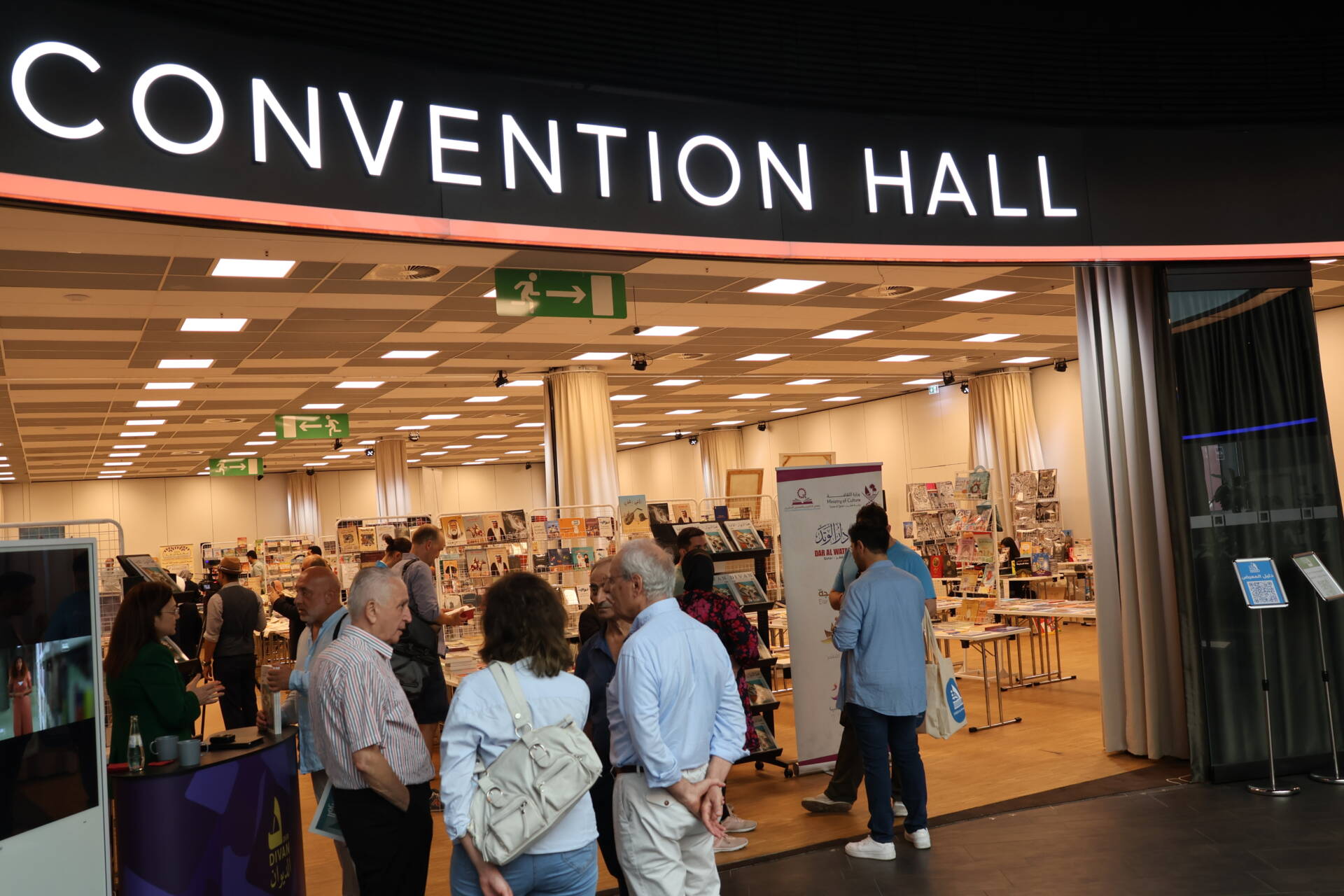 Eingangsbereich einer Convention Hall mit Messeständen, Besuchern, Informationsmaterialien und Vorhängen.