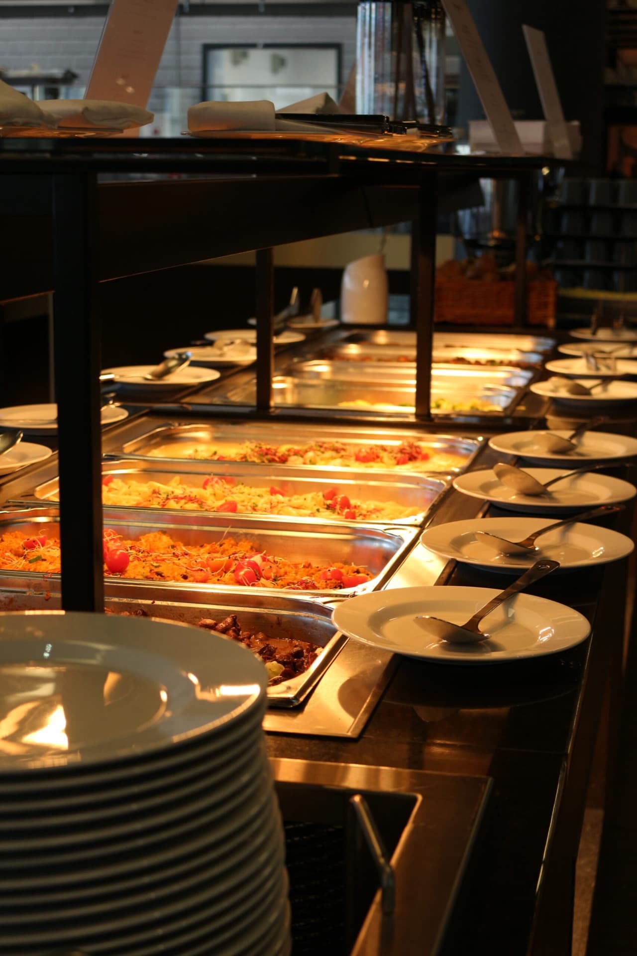 A buffet with several hot dishes in stainless steel containers and a stack of white plates with cutlery on the side.