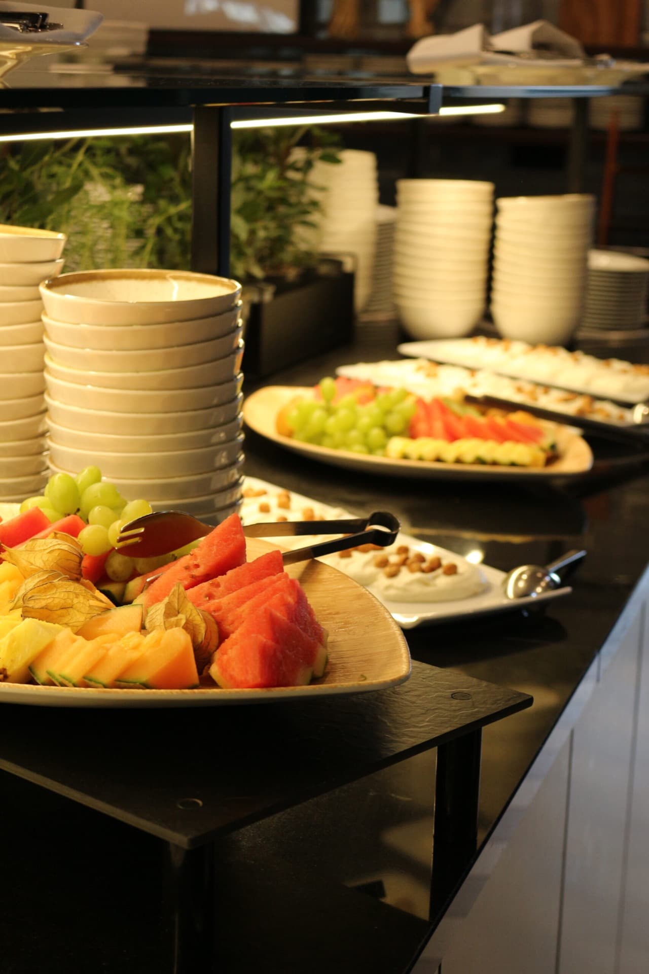 A buffet with plates and several platters of sliced fruit such as melon, pineapple and grapes on a black surface.